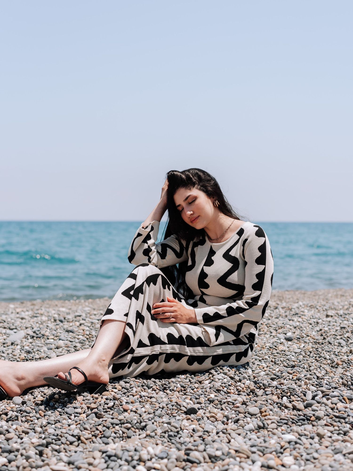Woman in black and white zigzag patterned dress sitting relaxed on rocky beach by the sea.