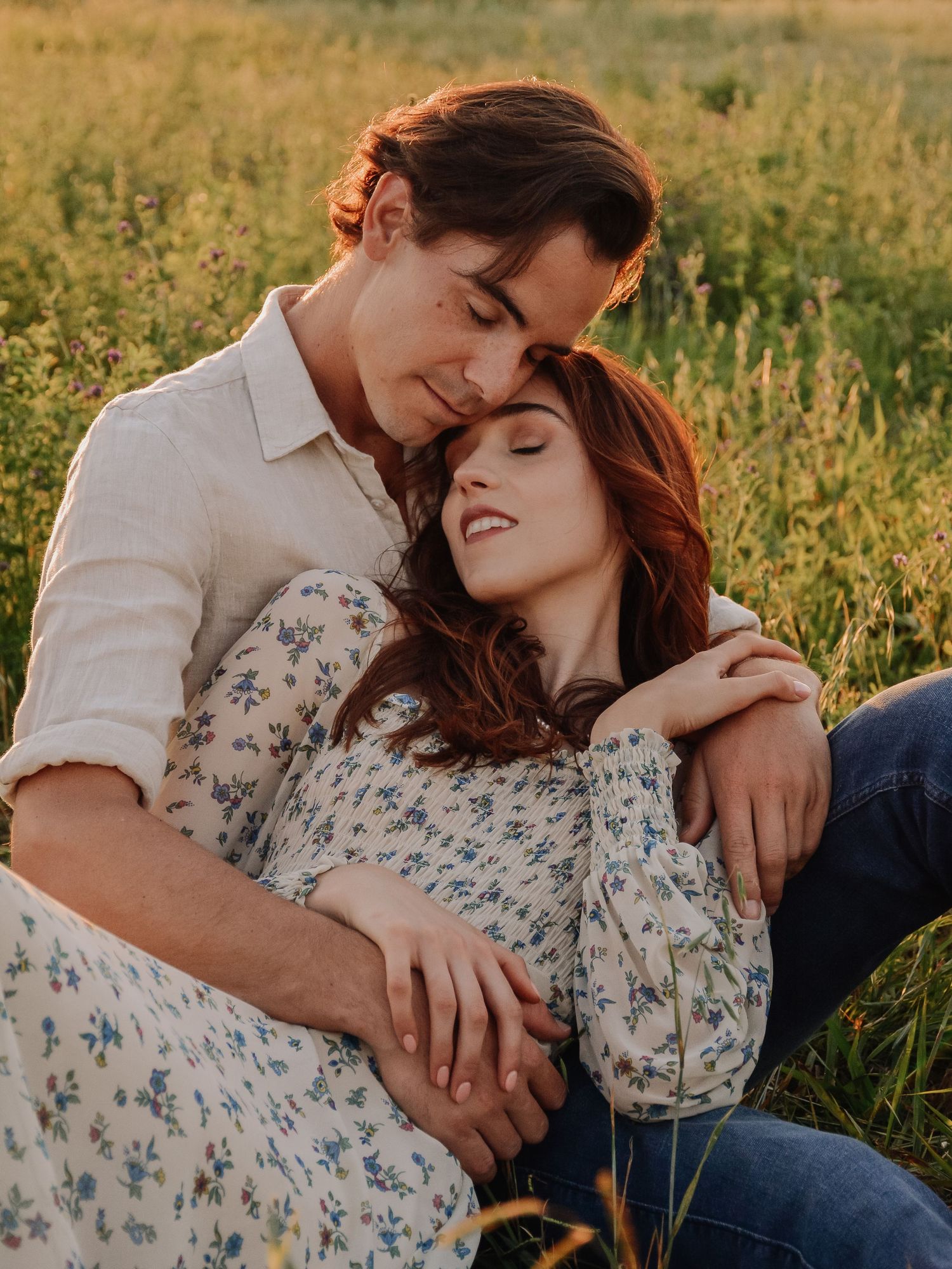 Couple embracing in a sunny green field, woman in floral dress leaning on man in white shirt during golden hour.