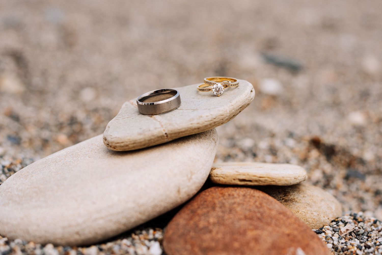 Two wedding rings, a gold diamond engagement ring and silver band, resting on smooth pebbles on a sandy beach.