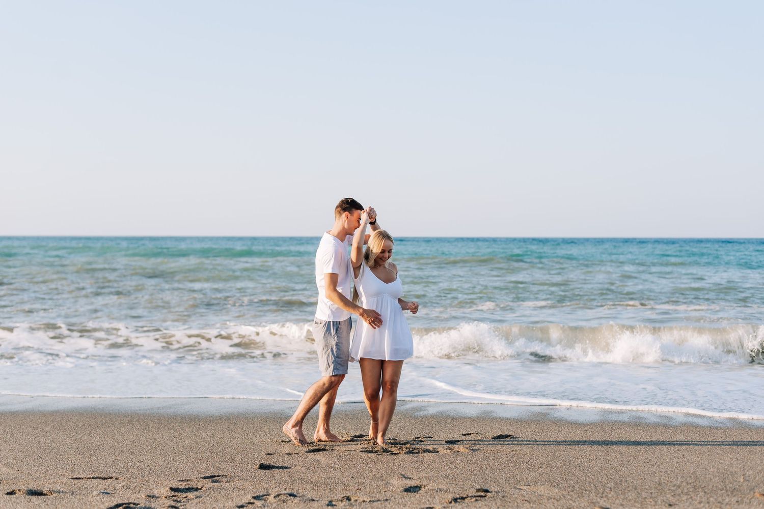 Couple in white outfits walking romantically on a sandy beach with ocean waves in the background.