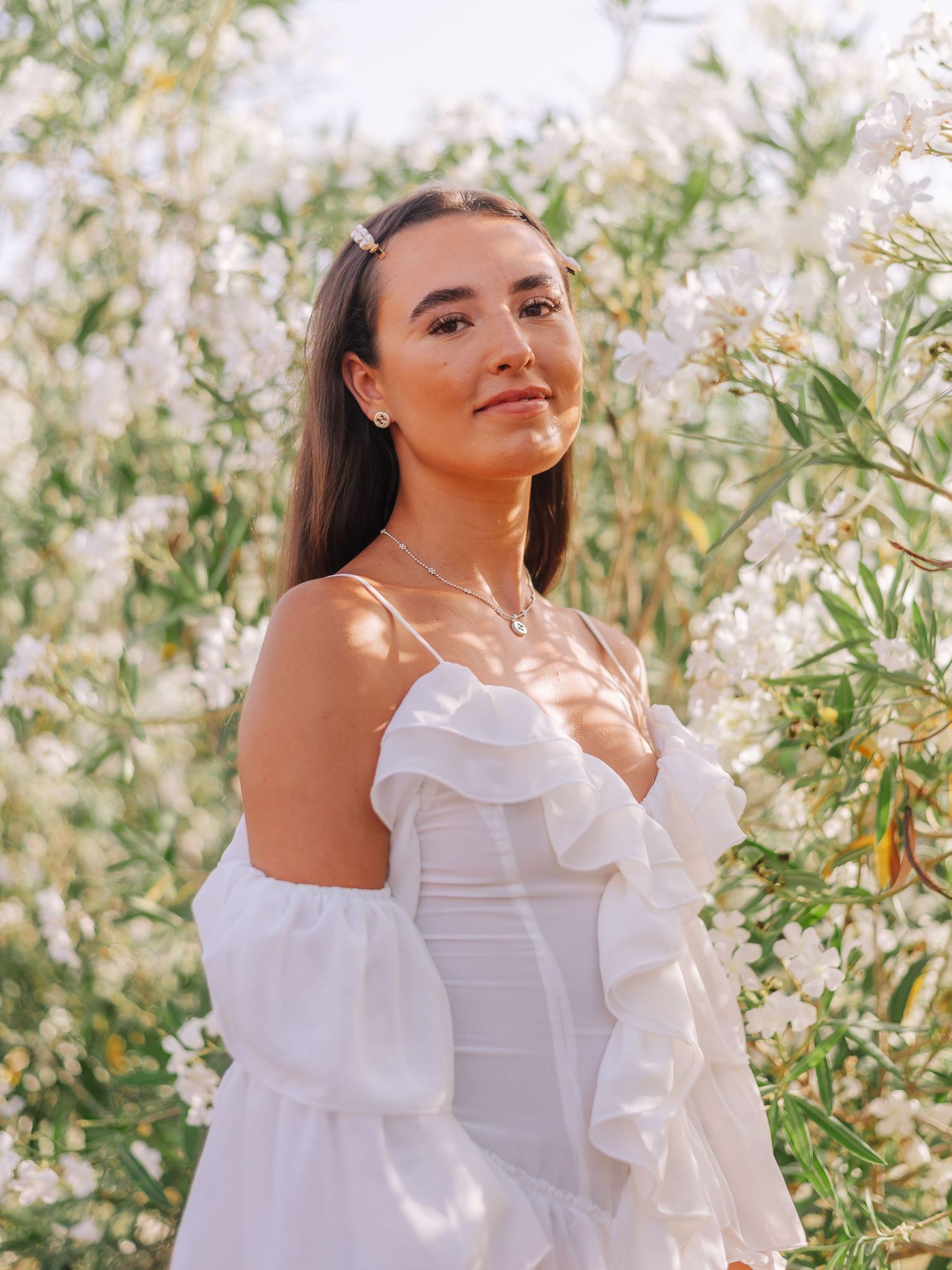 Young woman in white ruffled off-shoulder dress posing among blooming white flowers in sunlight.