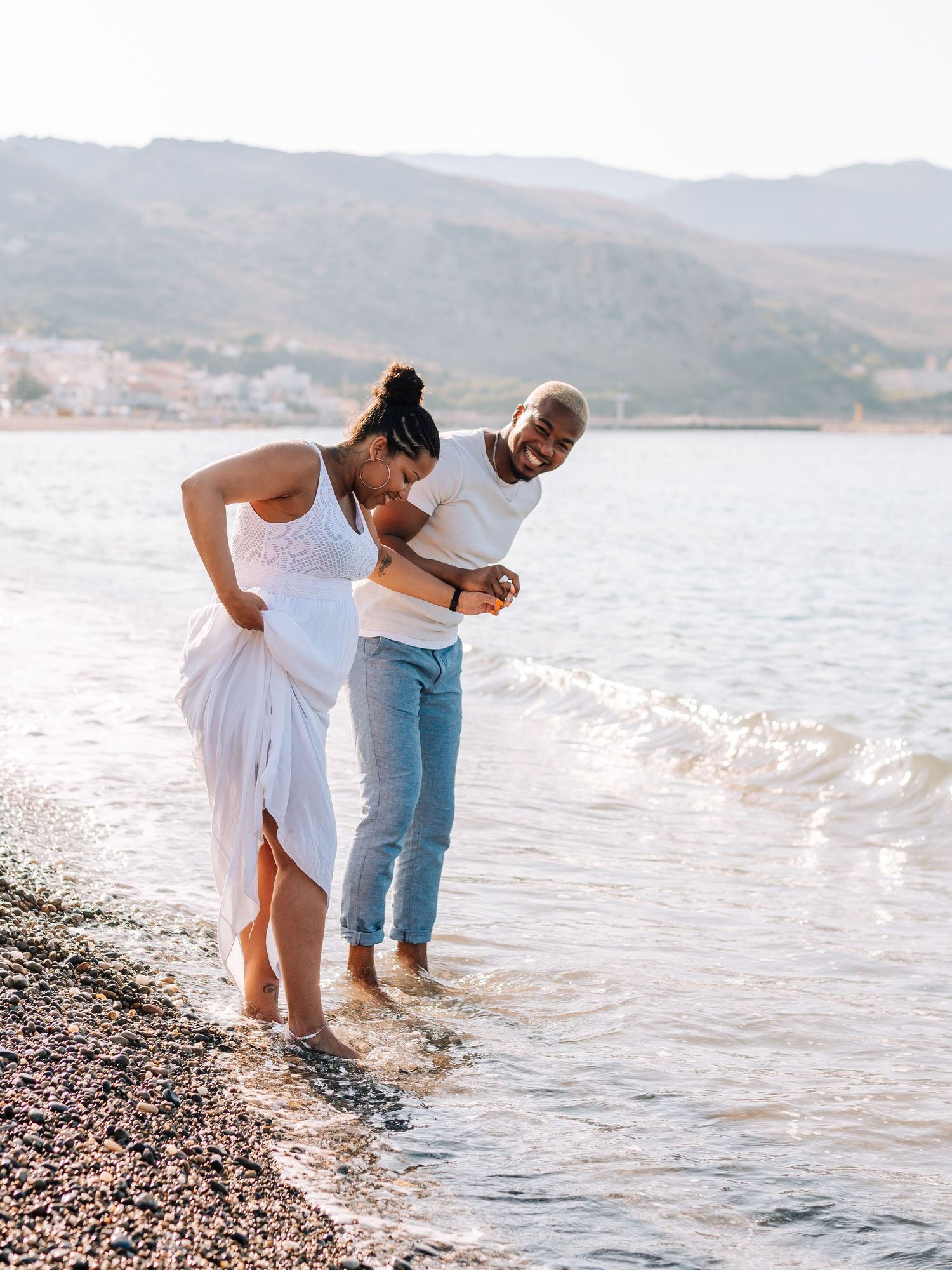 Pregnant couple laughing and wading in shallow beach water at golden hour with scenic mountain backdrop.