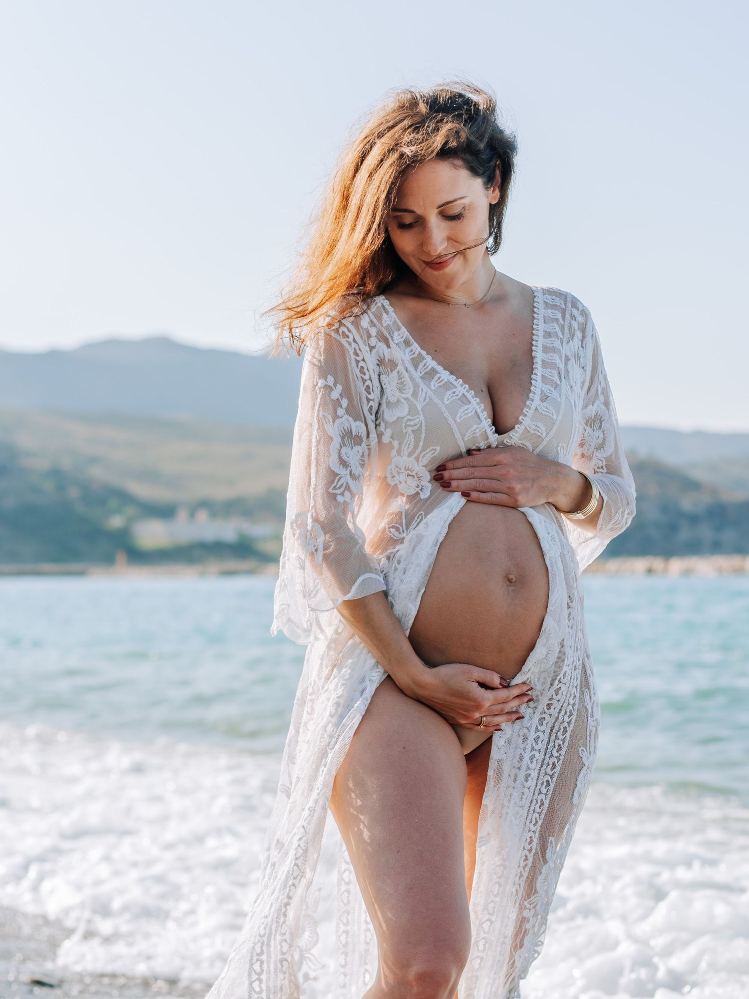 Pregnant woman in white lace kimono holding baby bump on sunny beach with mountains in the background.
