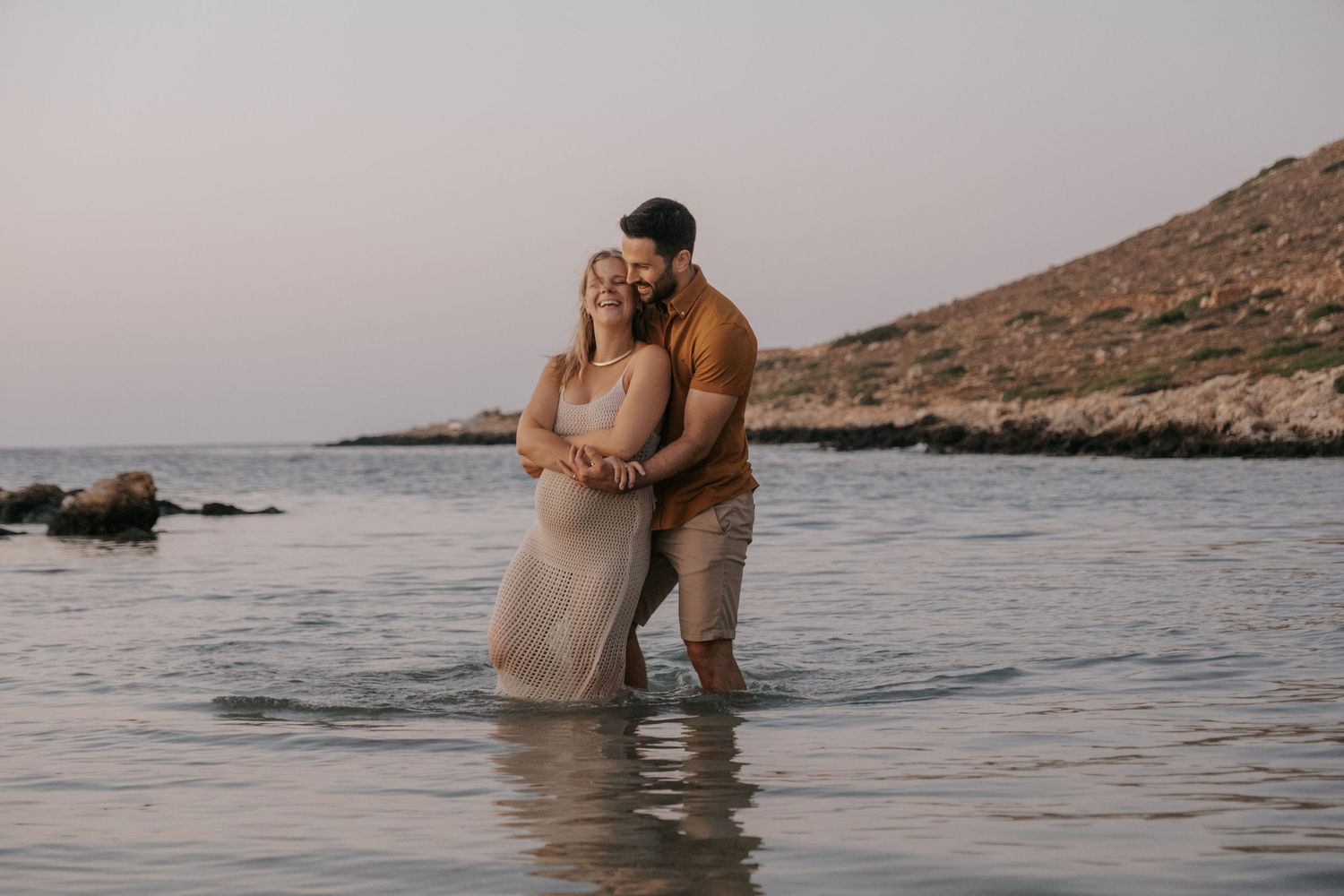 Romantic couple embracing while standing in shallow ocean water at a peaceful rocky beach.