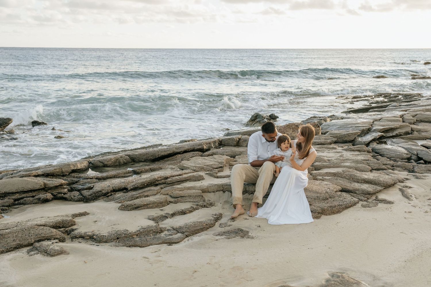 Couple sitting together on flat coastal rocks by the ocean shore in wedding attire.