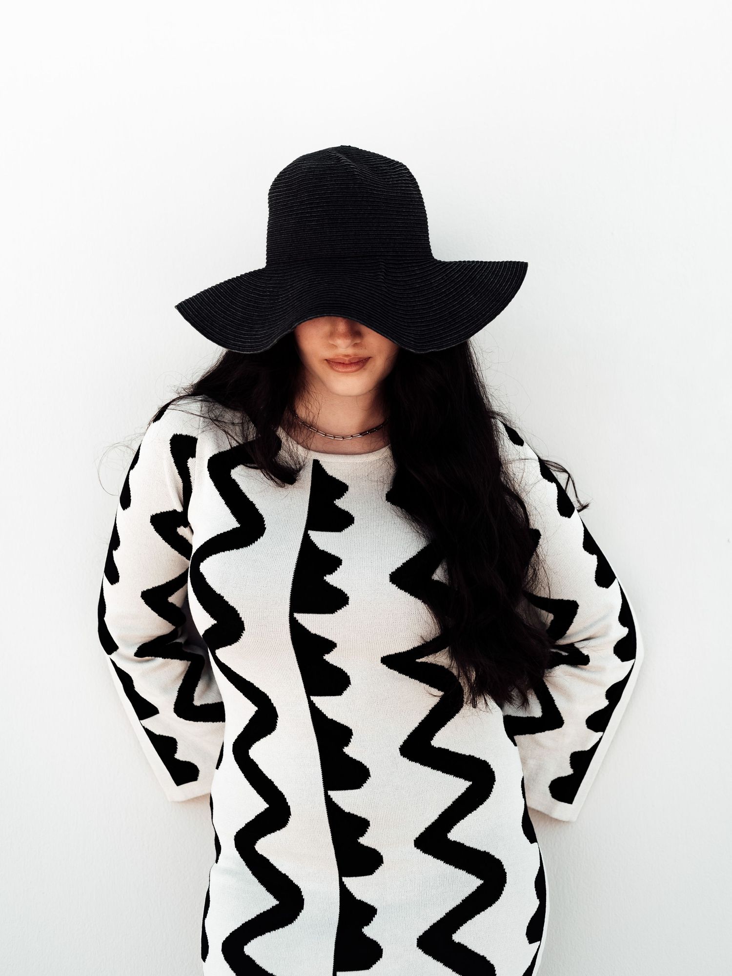 Woman in black and white zigzag knit dress wearing wide-brim black hat covering eyes against white wall.