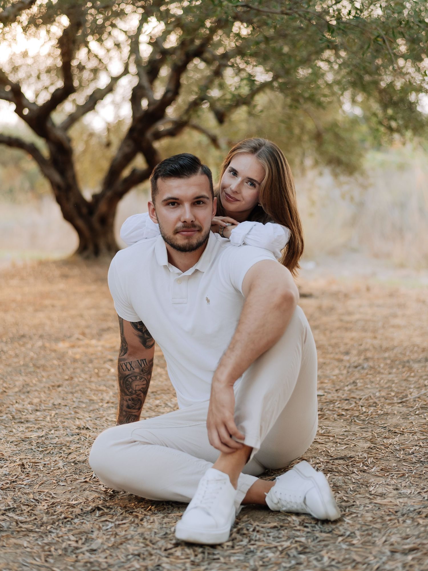 A couple in matching white outfits poses outdoors under a tree, man sitting cross-legged with woman leaning on his back.