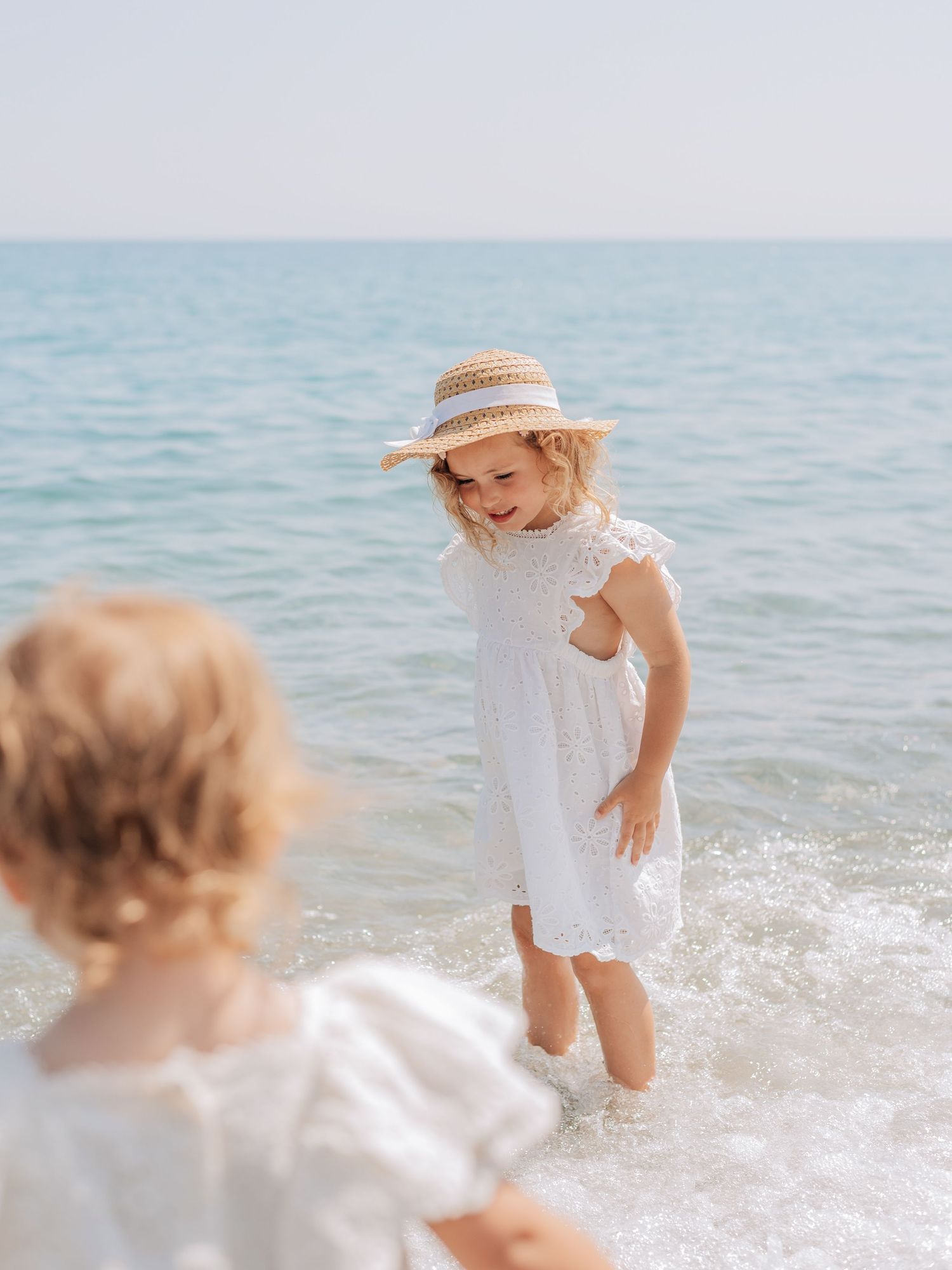 A smiling little girl in a white dress and straw hat plays in the ocean waves at the beach.
