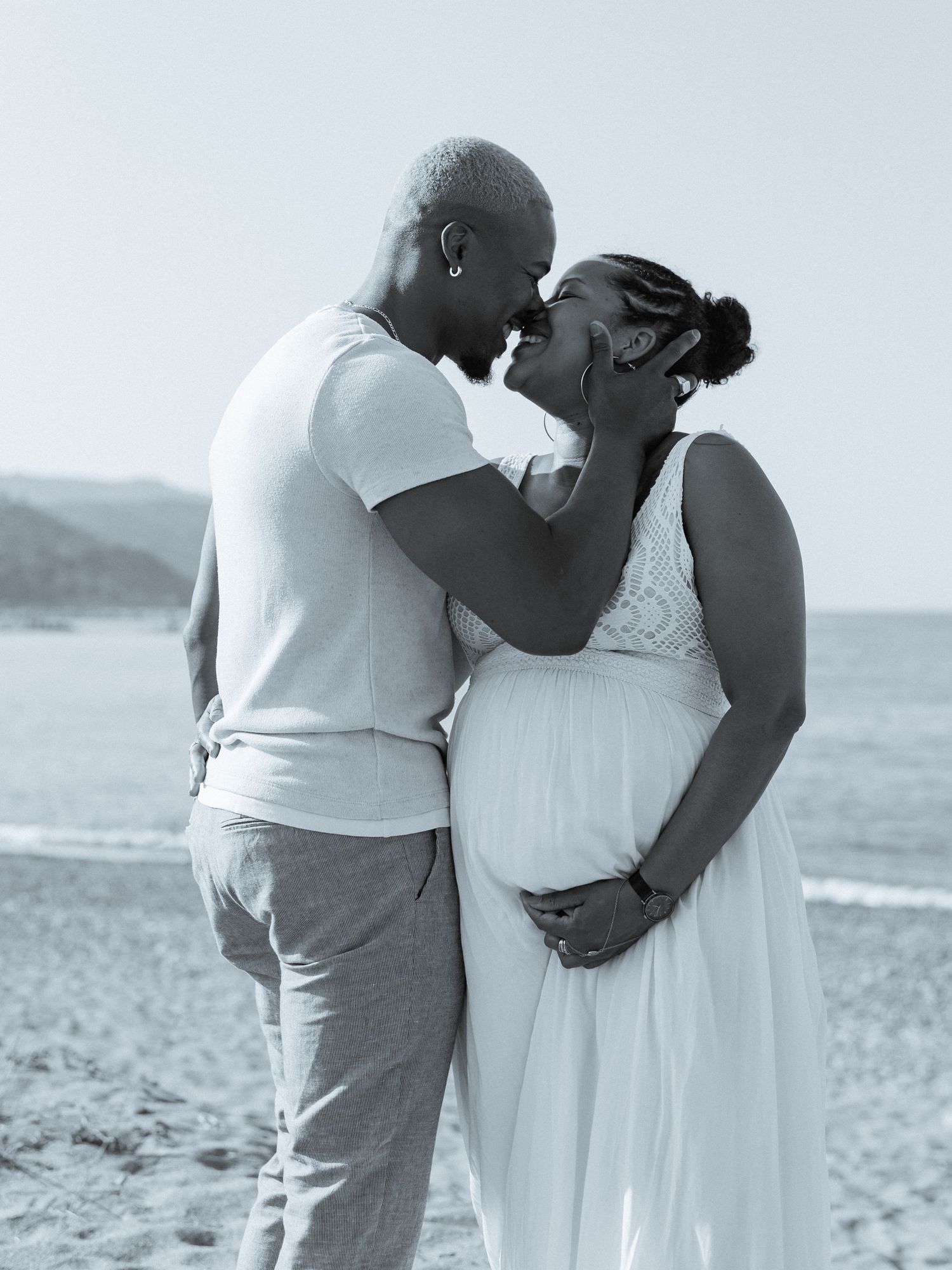 Black and white photo of a pregnant couple sharing a tender moment on a beach, the man gently holding her face.