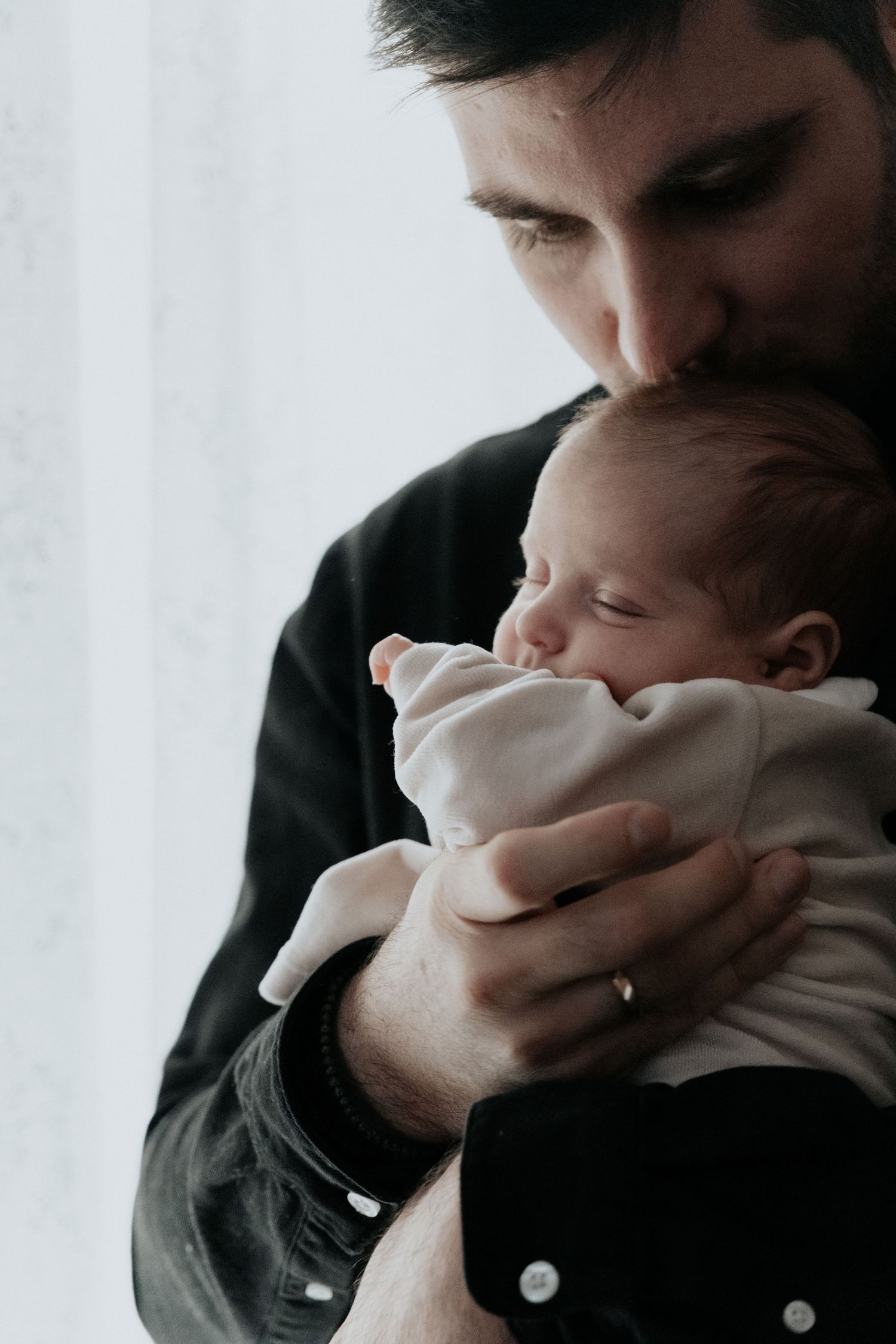 A father tenderly kisses his sleeping newborn baby while cradling the infant in his hands.