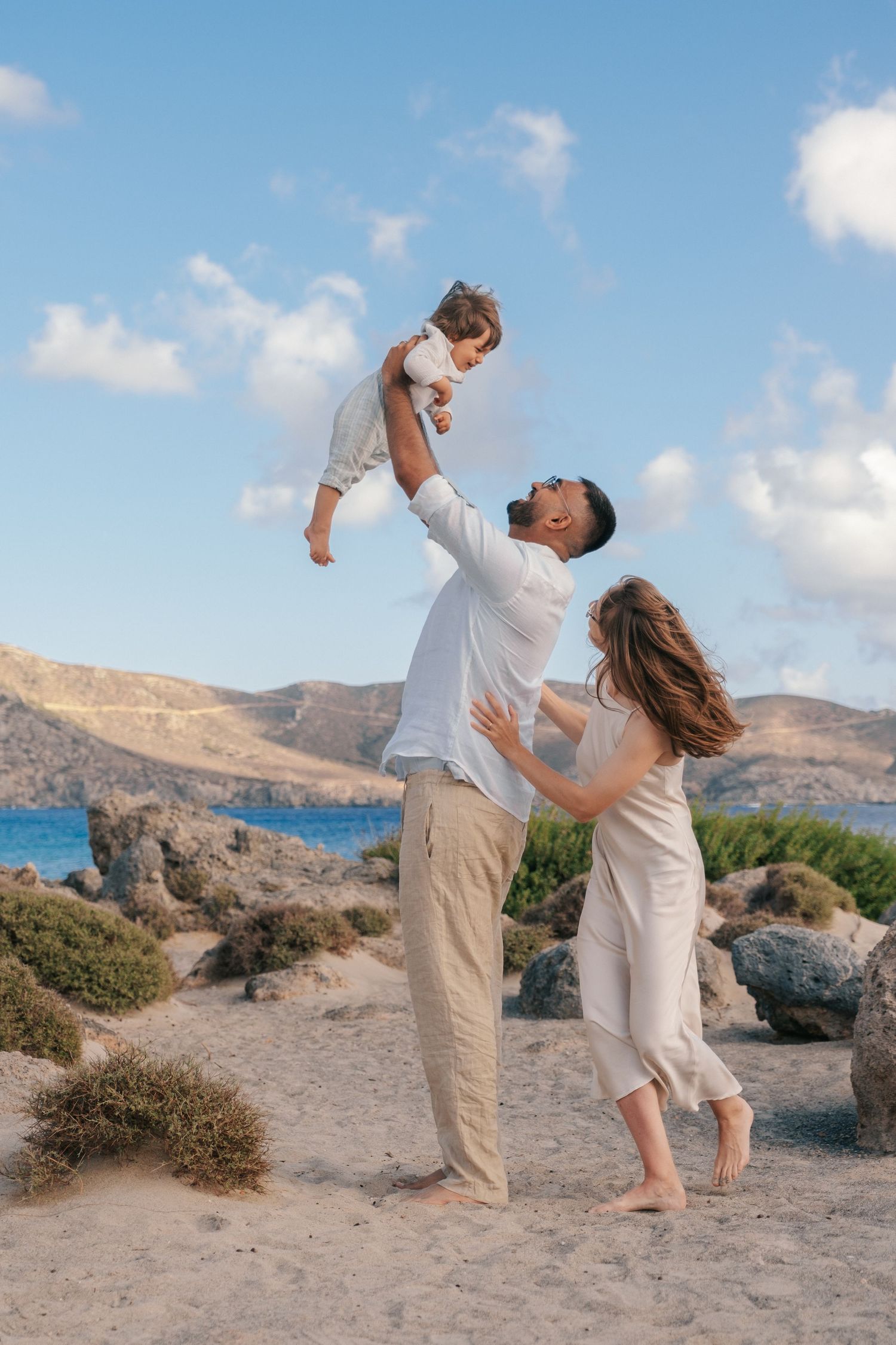 Father lifting baby in air while mother watches on rocky Mediterranean beach.