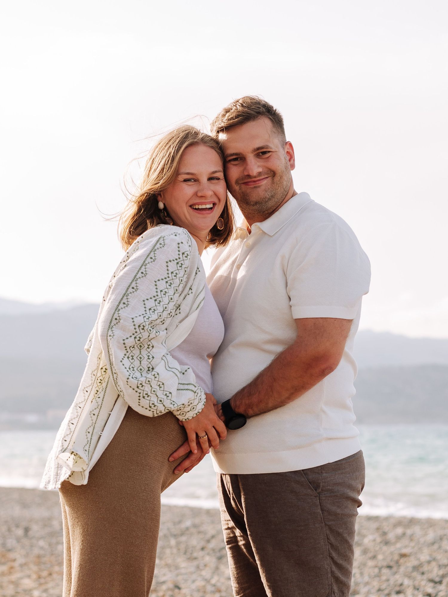 Smiling couple embracing on a rocky beach with mountains and ocean in the background.