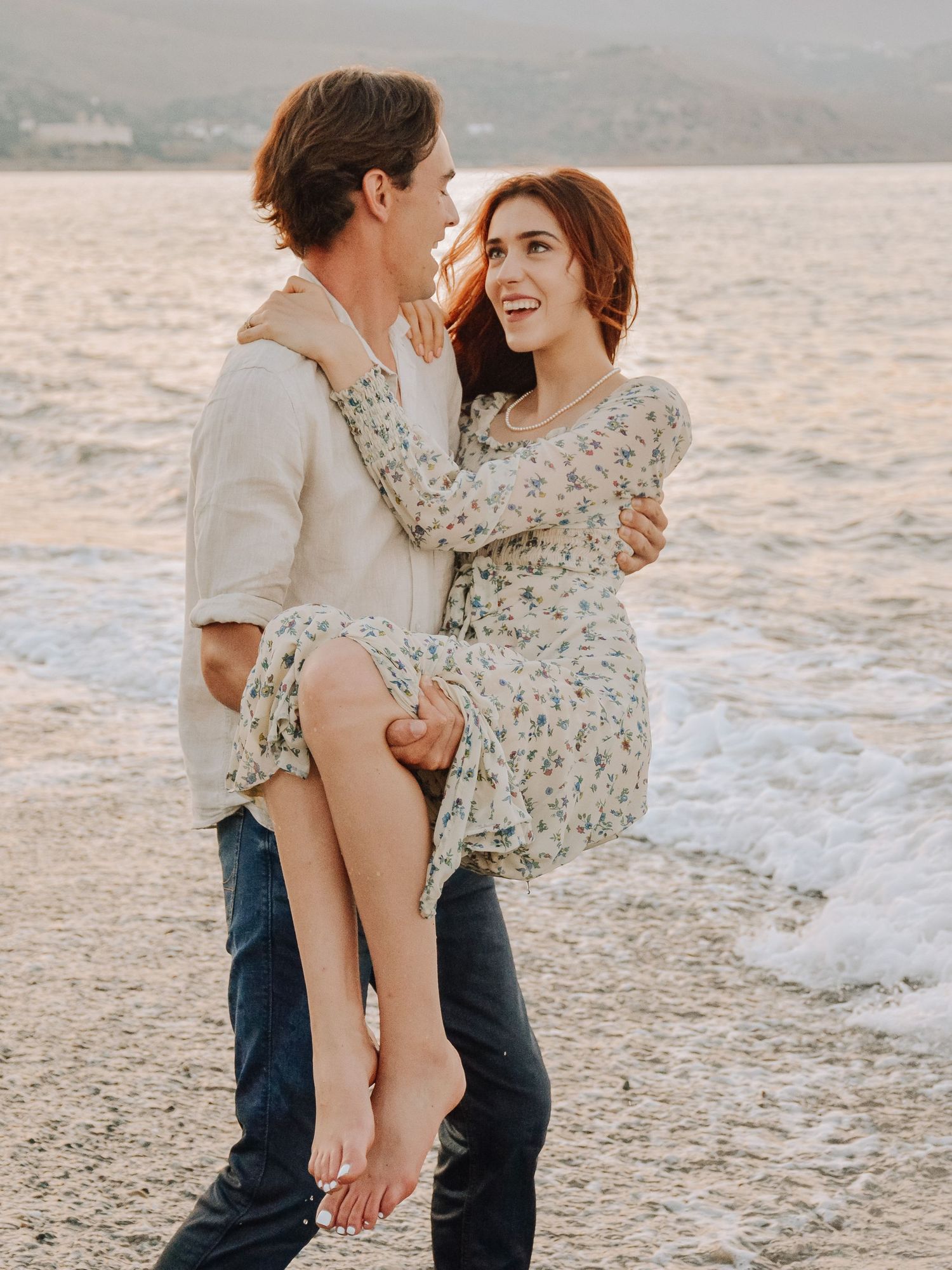 A couple shares a romantic beach moment, man lifting woman in floral dress, waves crashing behind them at sunset.