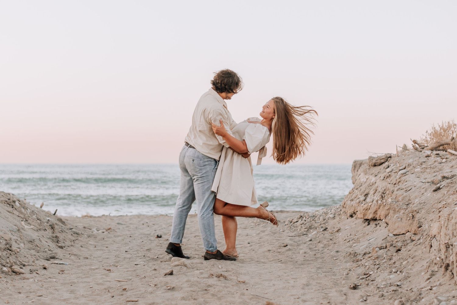 Couple dancing romantically on a sandy beach at sunset with ocean waves in the background.