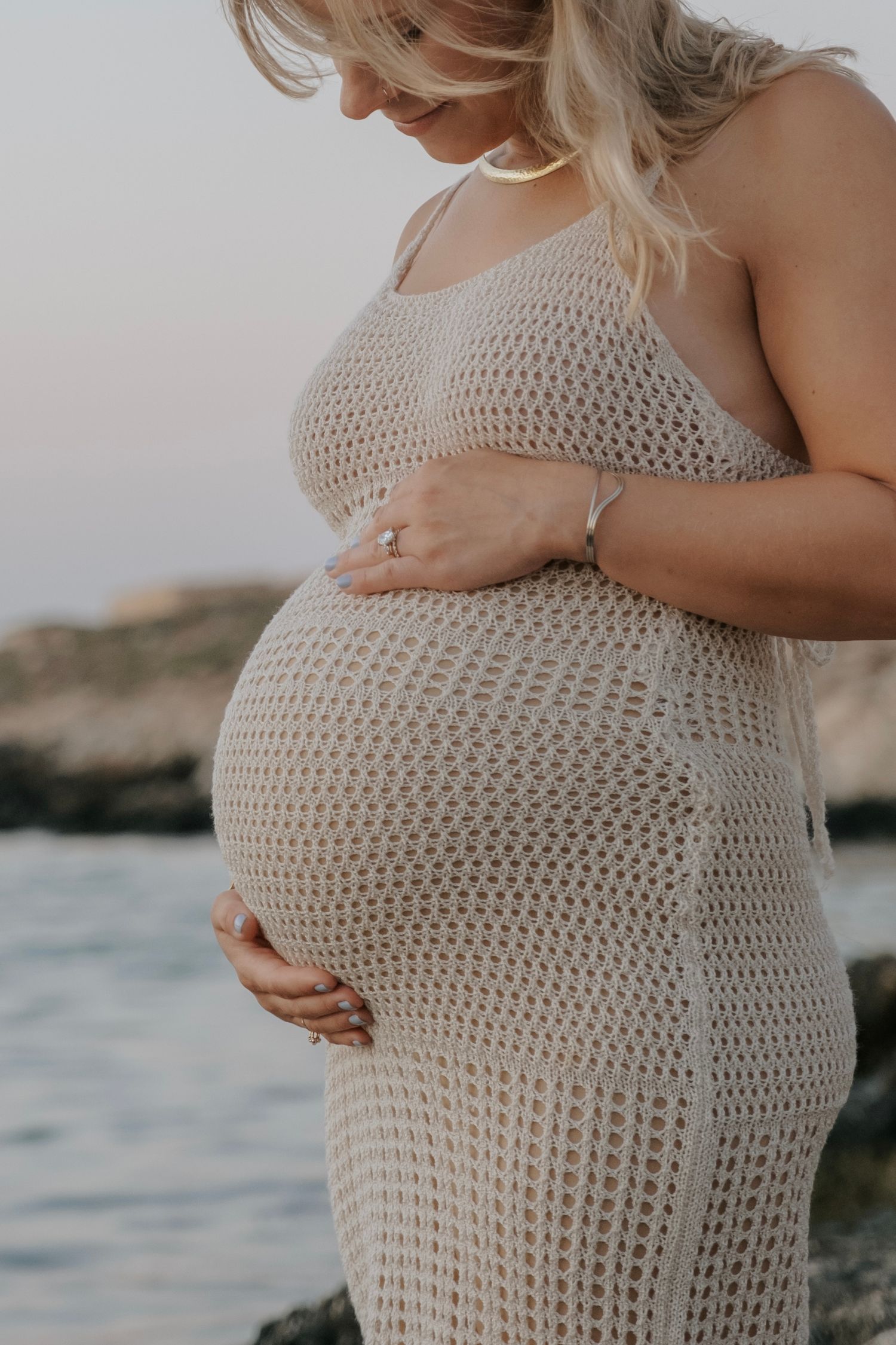 Close-up of pregnant woman in beige crochet dress gently holding her baby bump near the sea.