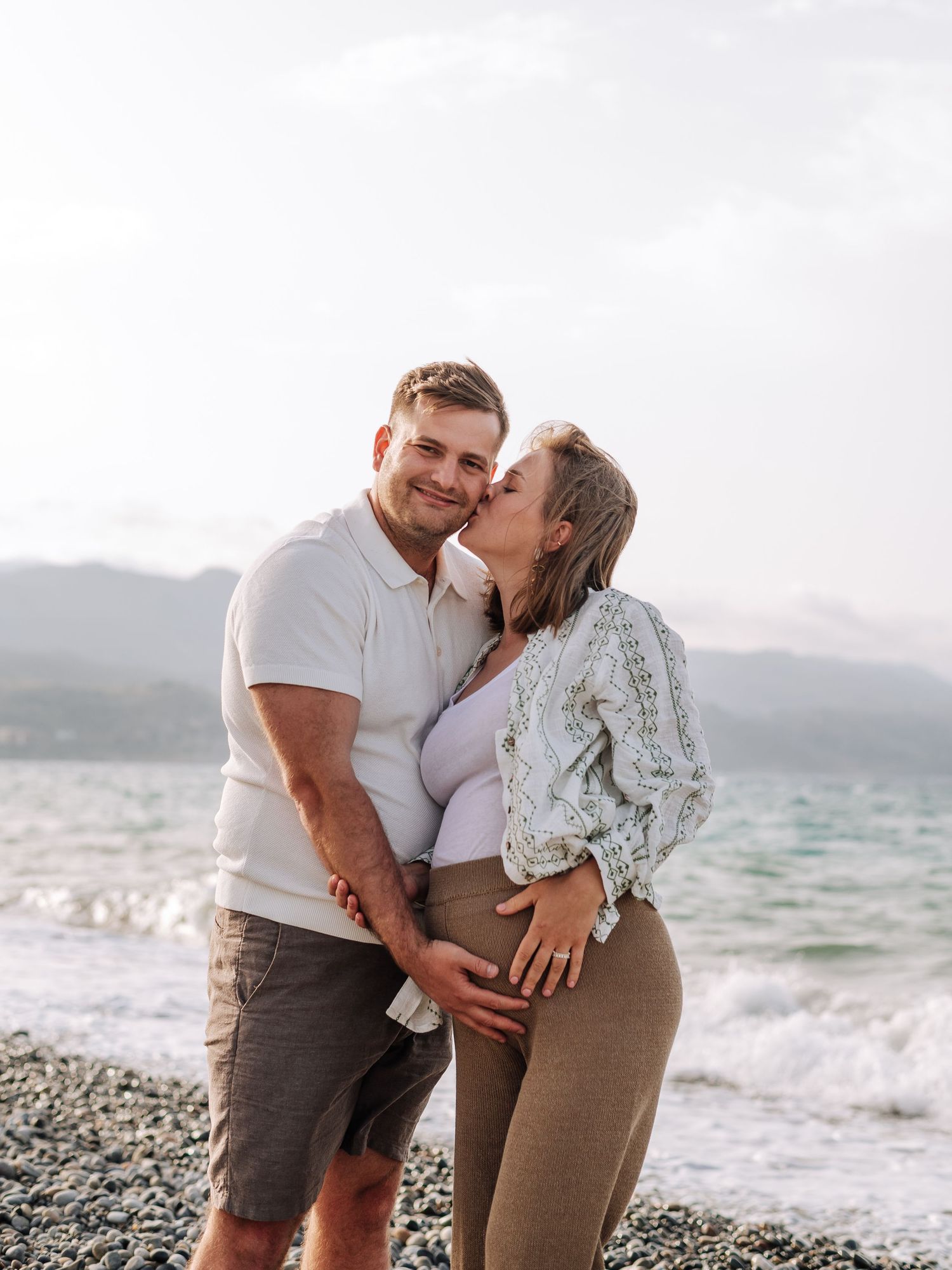 Couple sharing a kiss on rocky beach, woman visibly pregnant in white cardigan and tan pants.