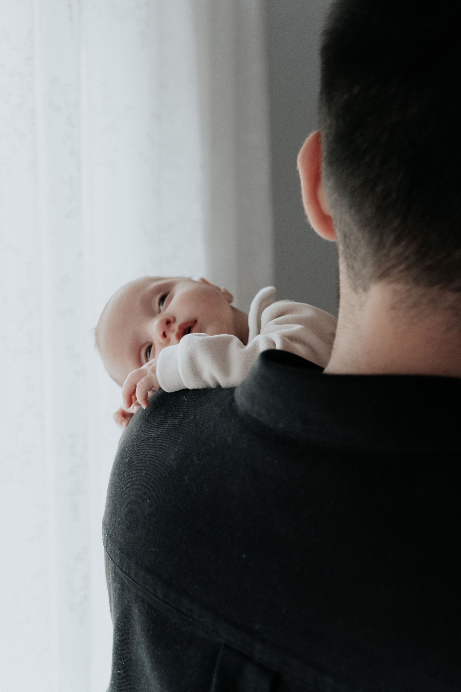 Father in black sweater holding newborn baby against his shoulder near a bright window.