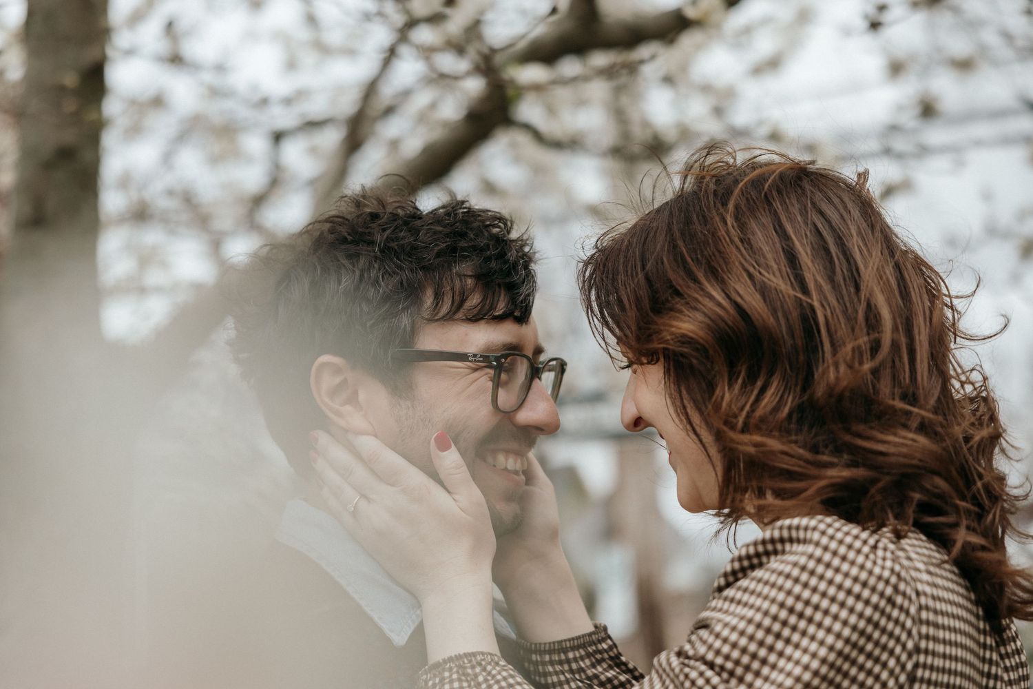 A couple smiles at each other outdoors, the woman gently cupping the mans face near bare winter trees.
