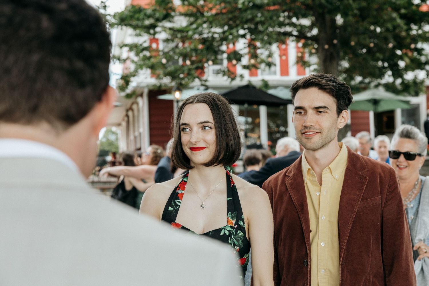 A woman in a floral halter dress and a man in a brown jacket chat with a guest at an outdoor wedding reception.