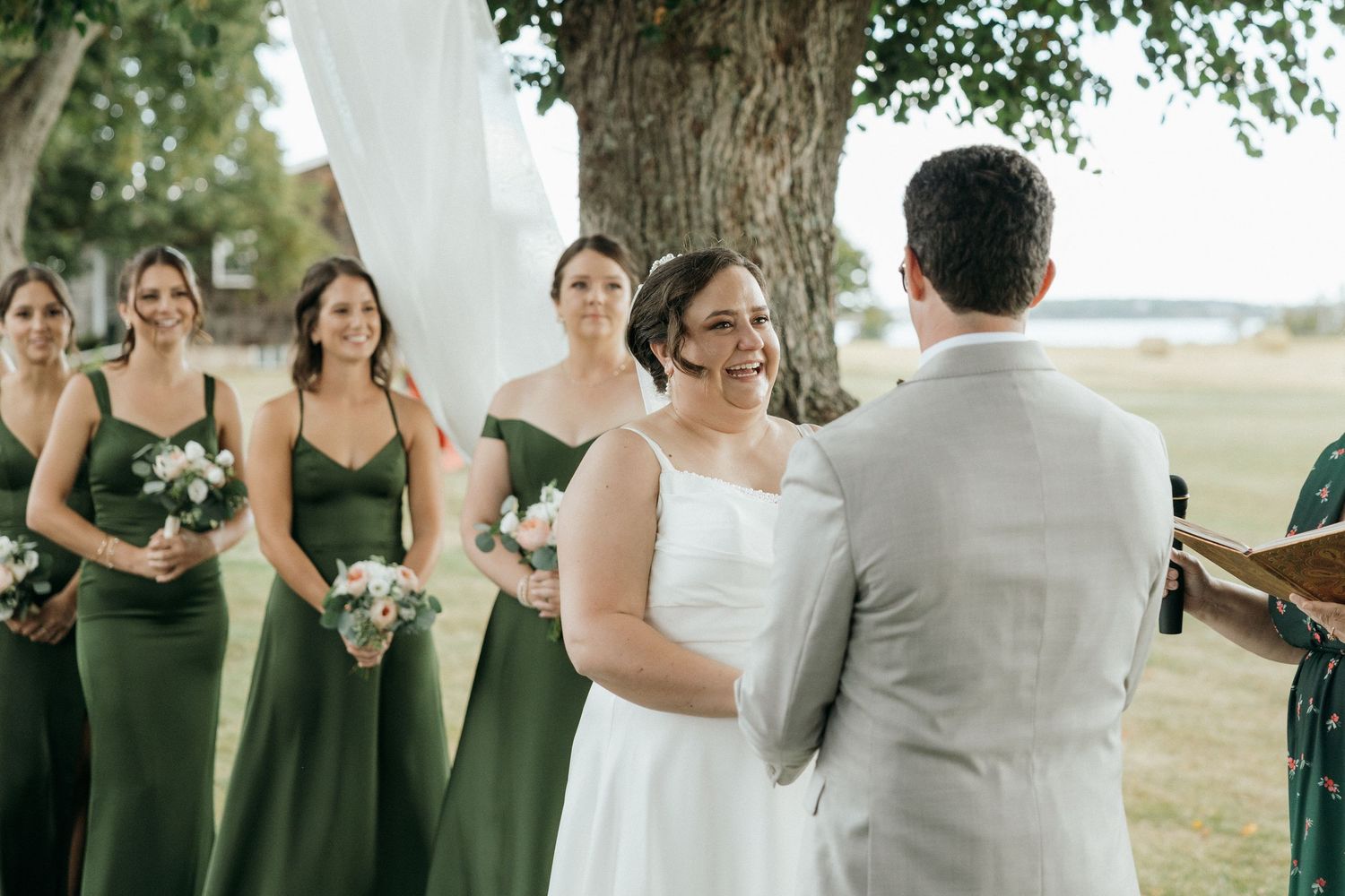 Bride in white gown smiles at groom during outdoor ceremony with green-dressed bridesmaids behind her.