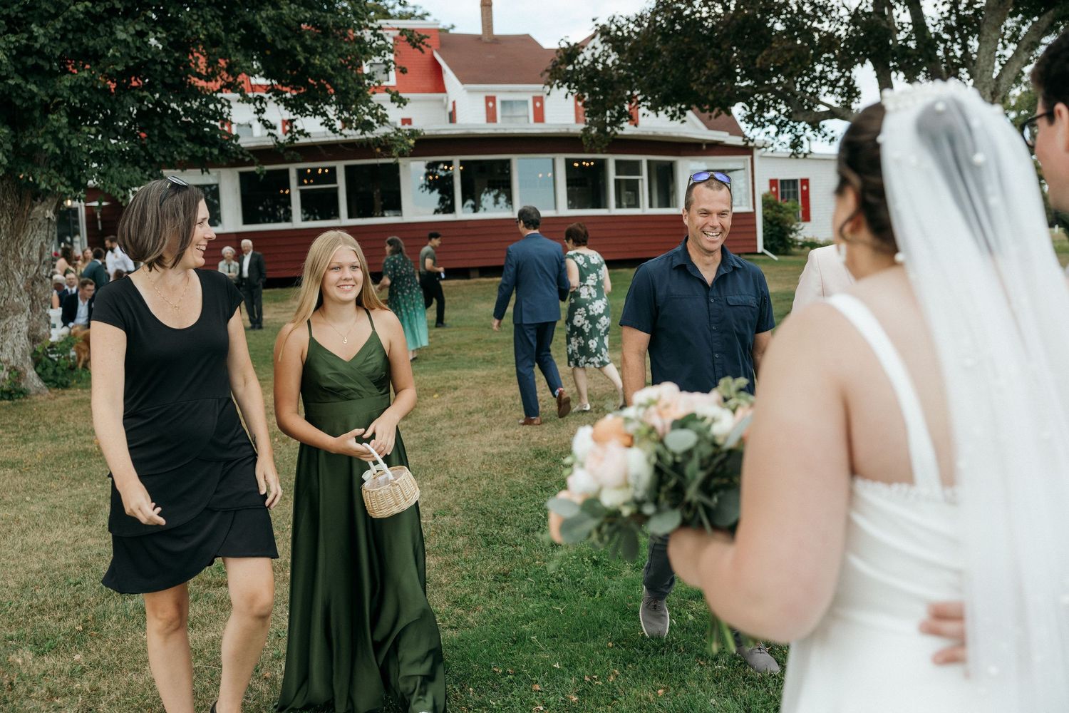 Bride in white gown holds bouquet while guests in green and black dresses approach outdoors.