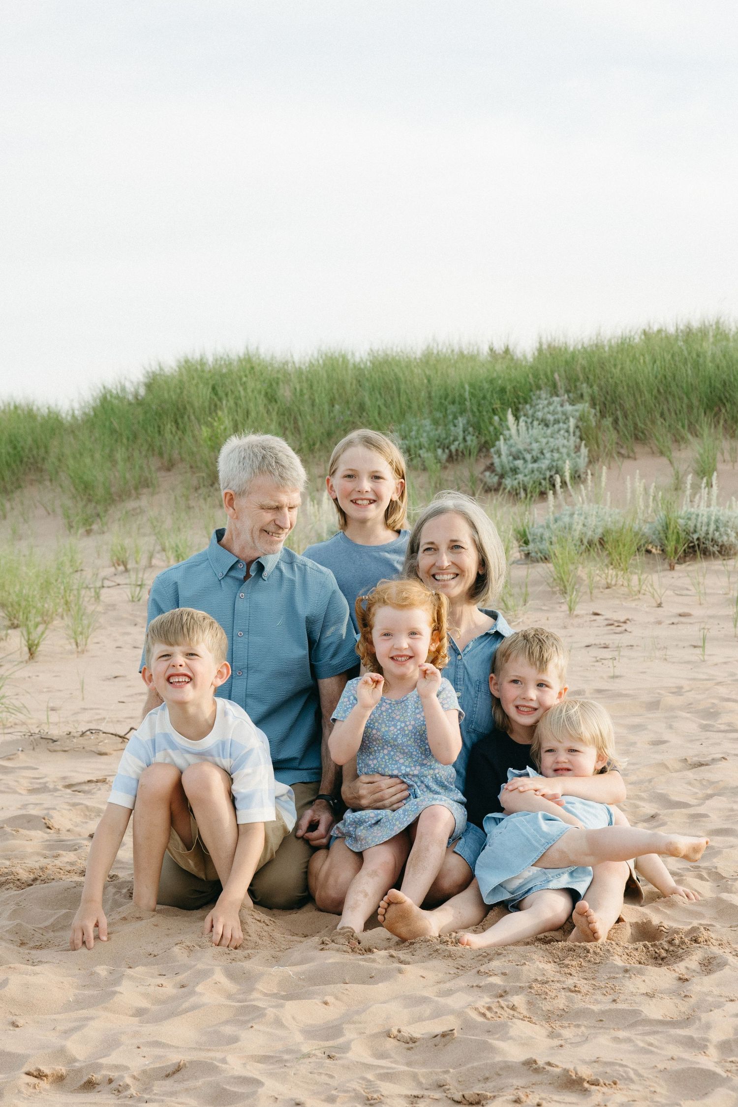 Grandparents and four young grandchildren smile together on sandy beach dunes during a family portrait session.