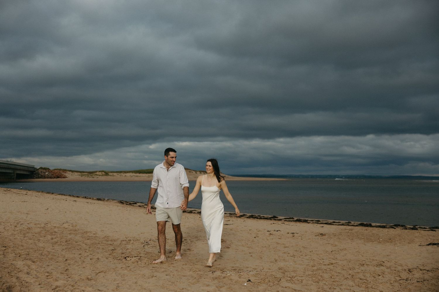 A couple in white outfits walking along a dramatic beach shoreline under moody storm clouds.
