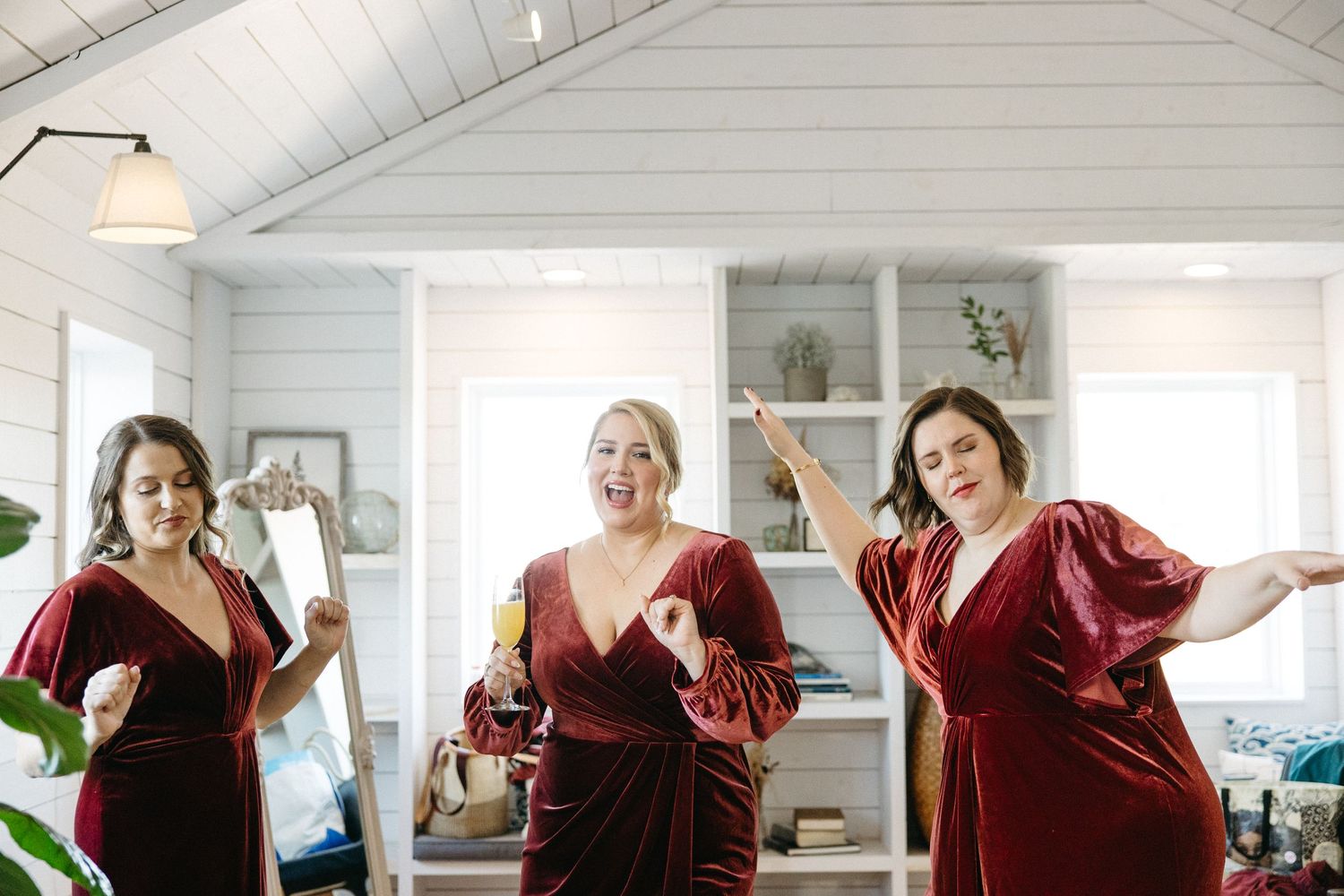 Three bridesmaids in burgundy velvet dresses pose together in a bright white room.