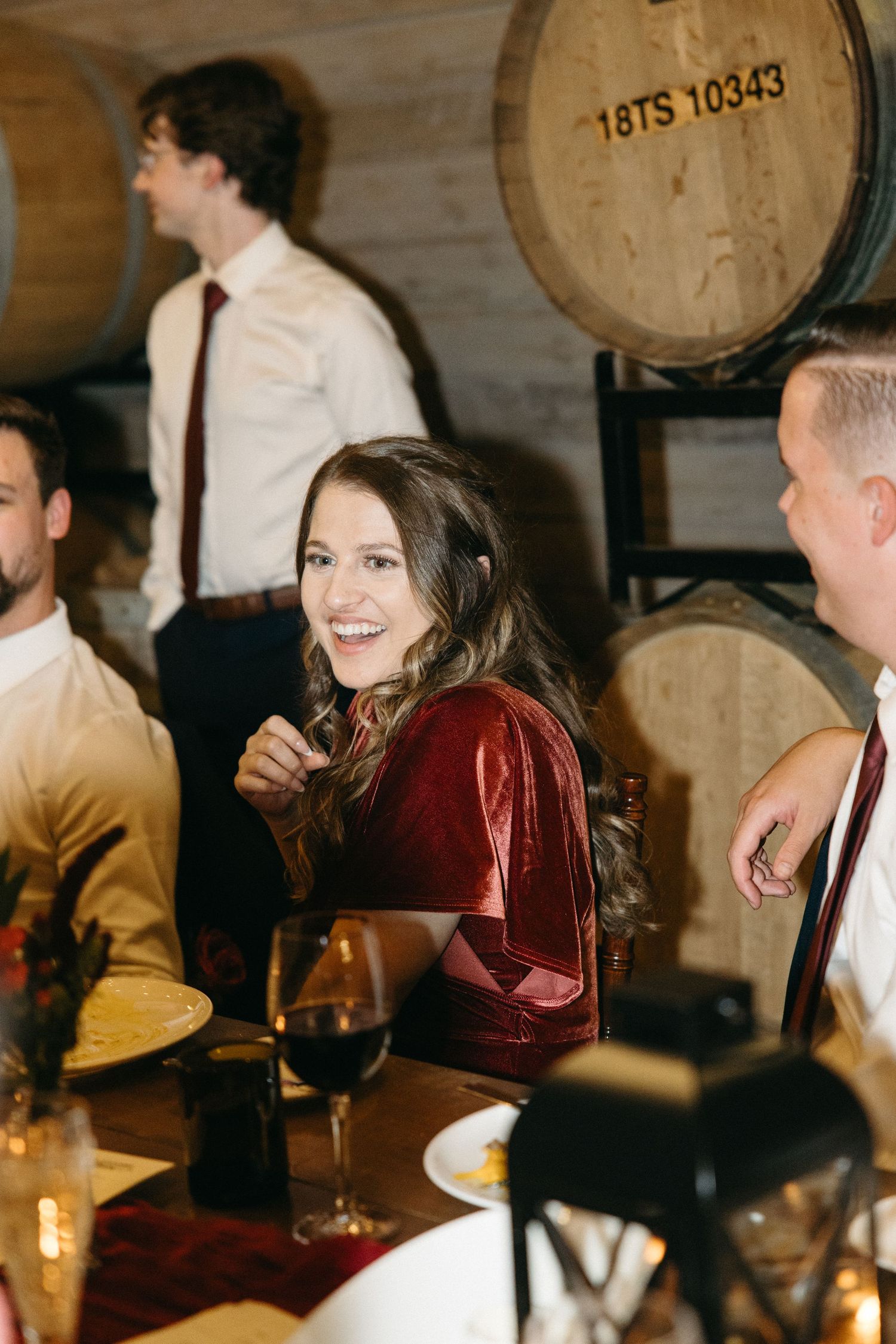 Guests enjoying dinner and conversation in a wine cellar with wooden barrels in the background.