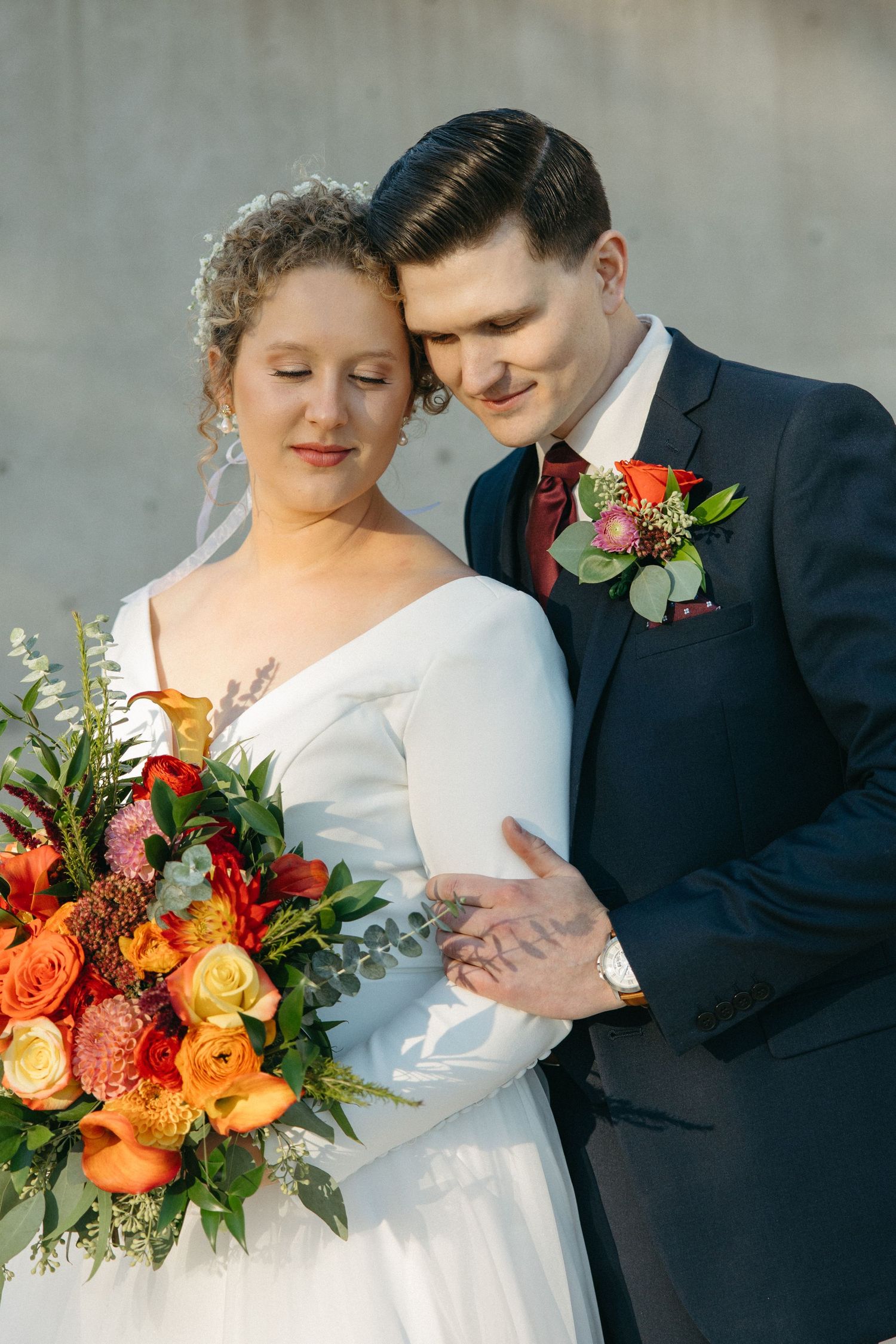 Wedding couple poses with vibrant orange and red floral bouquet against concrete wall.