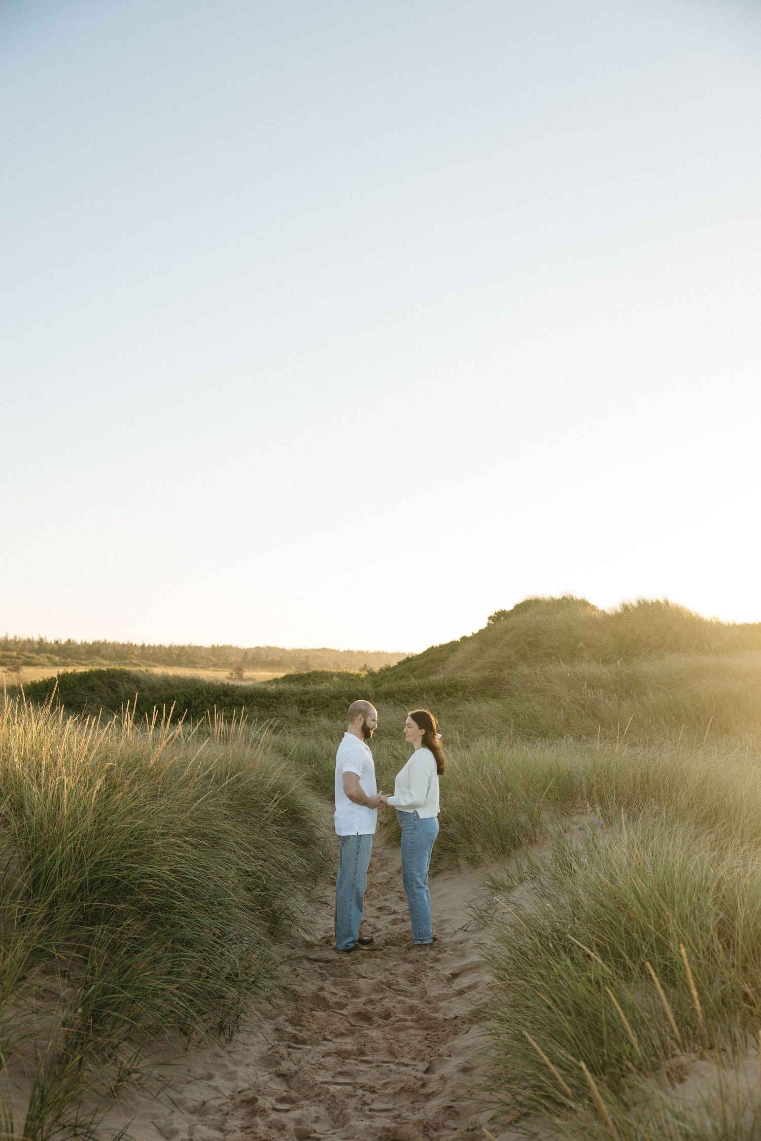 Two people standing together on sandy dunes with tall grass at golden hour.