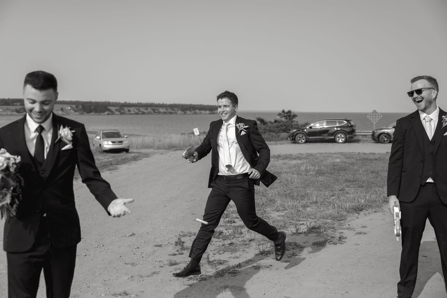 Three formally dressed attendees strike playful poses on a coastal road.