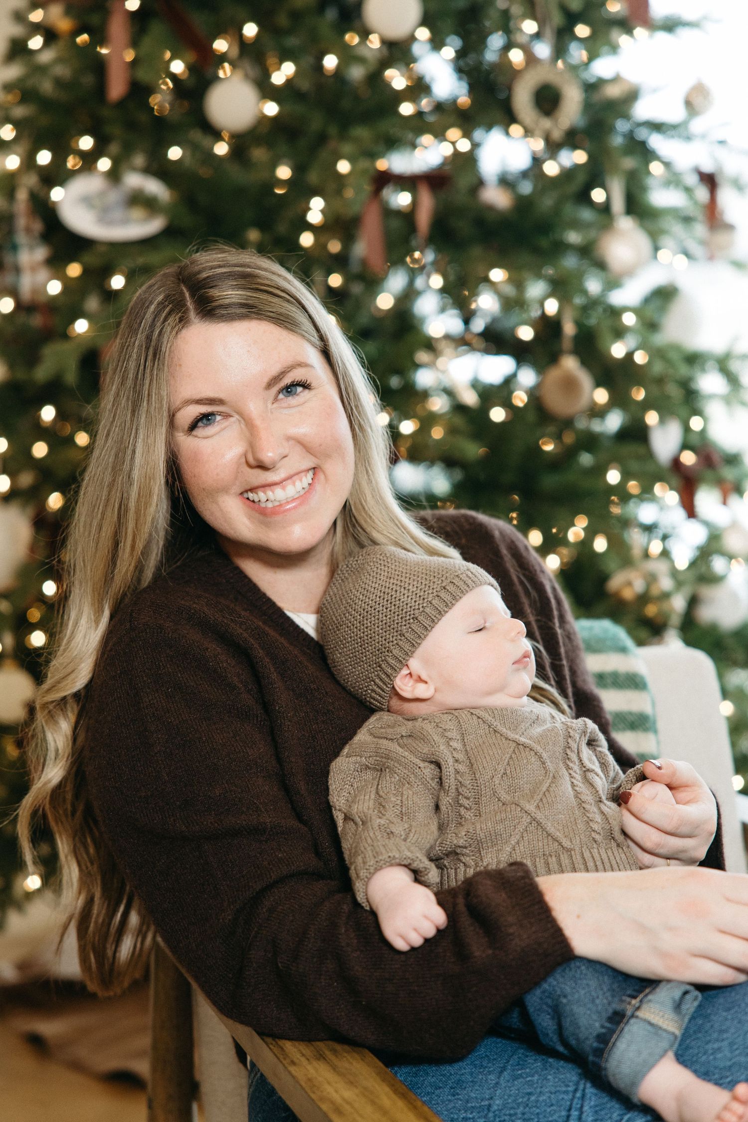 Woman in dark sweater holding baby in brown knit sweater near Christmas tree with twinkling lights.