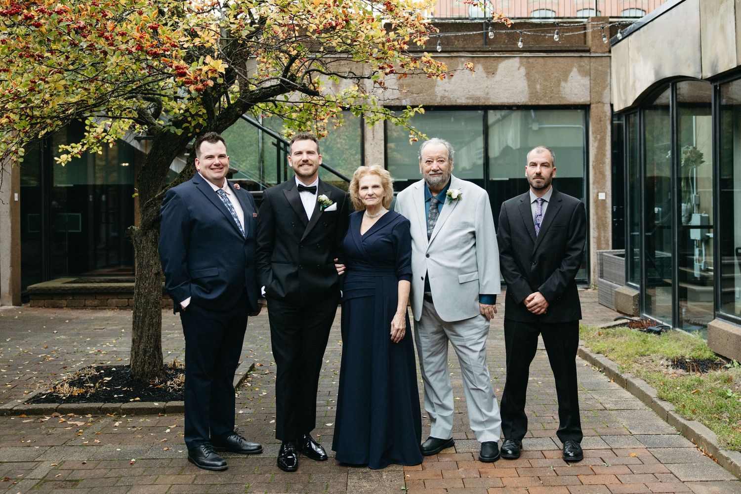A formal group photo of five people in evening attire standing under a tree with autumn leaves.