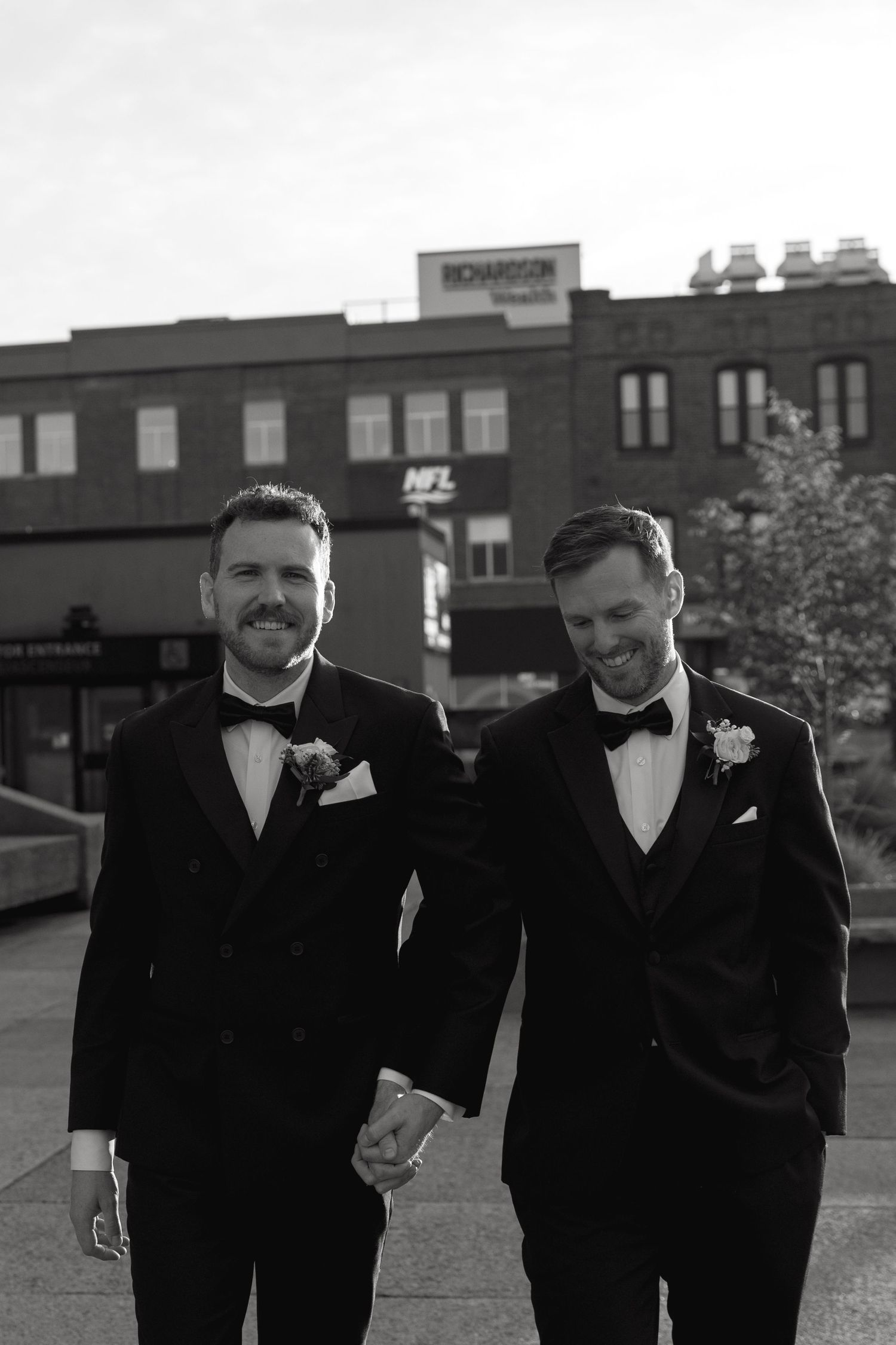 Two people in formal black tuxedos walking and laughing together outdoors.