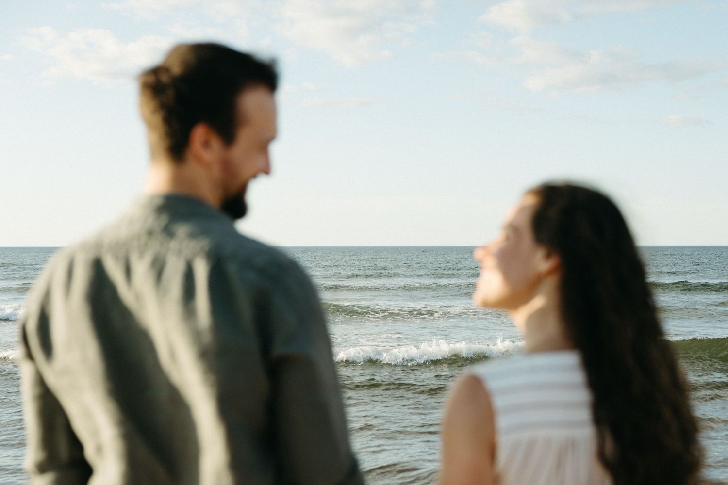 A couple smiles at each other on a beach with ocean waves in the background on a sunny day.