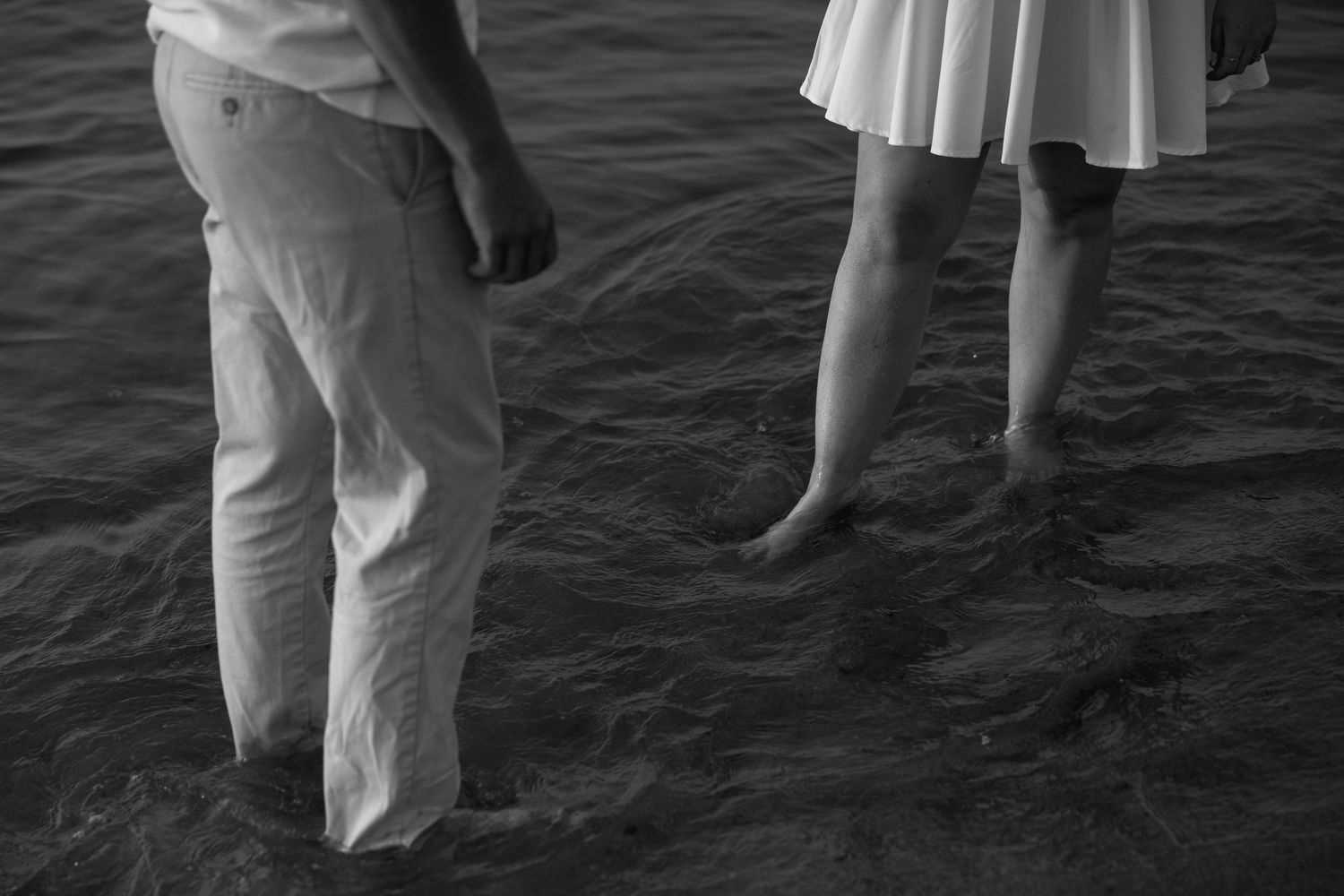 Black and white photo of a couple holding hands while standing barefoot in shallow ocean water.