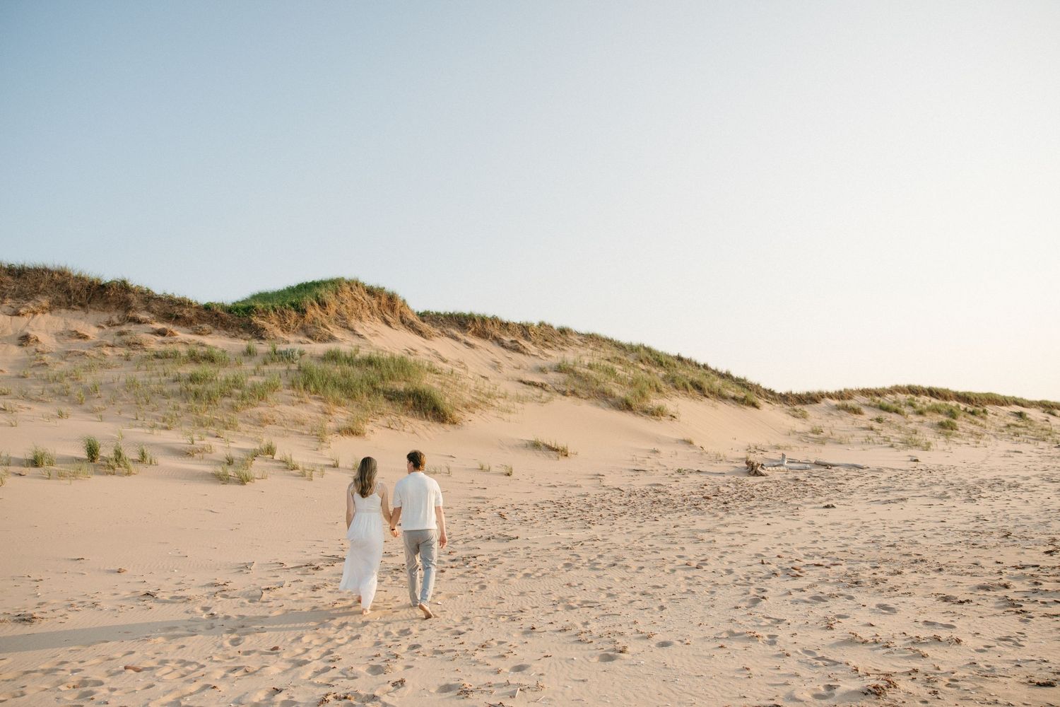 Couple walking hand in hand on sandy beach dunes under clear blue sky.