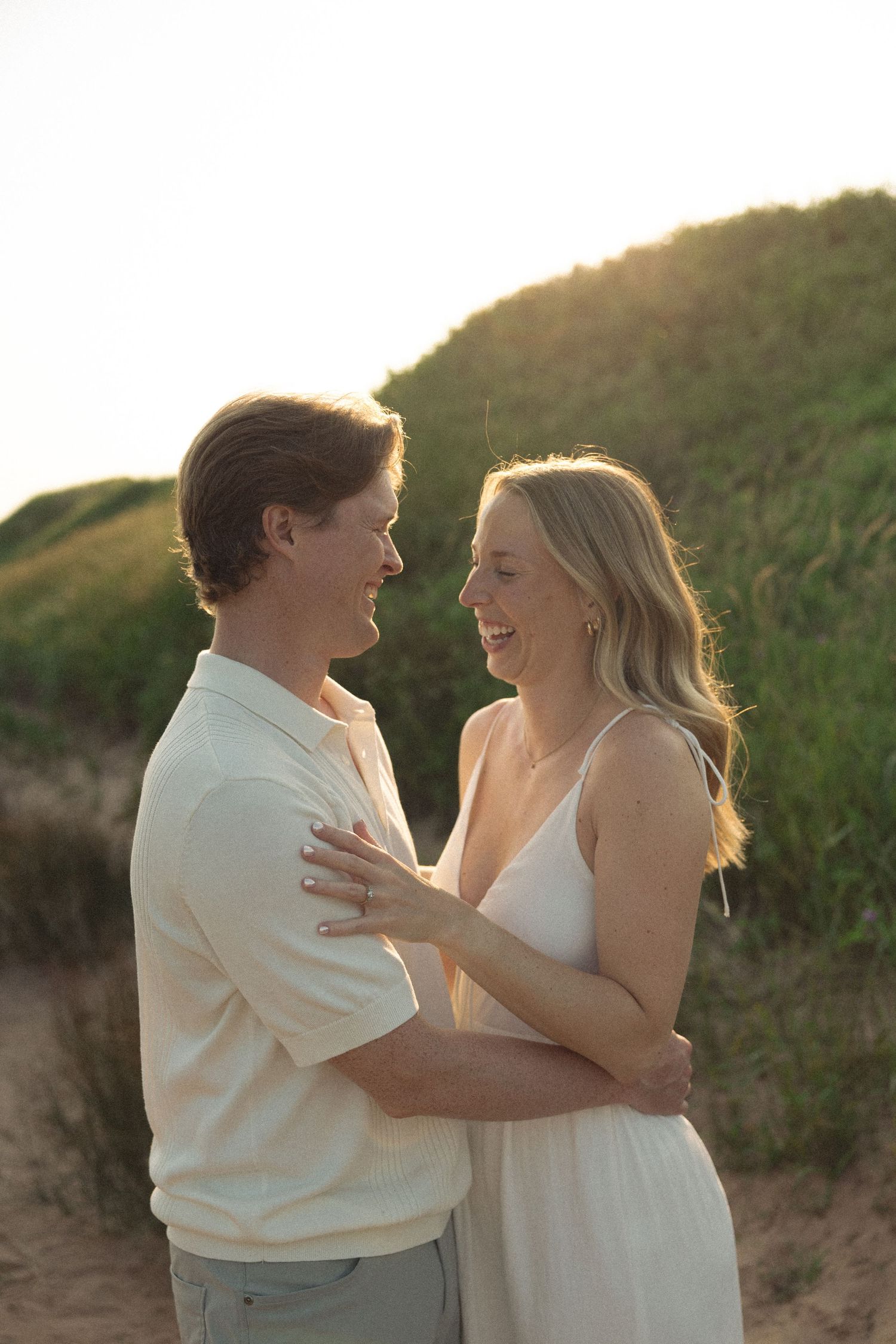 Couple embracing and laughing together at golden hour on a beach with green sand dunes in background.