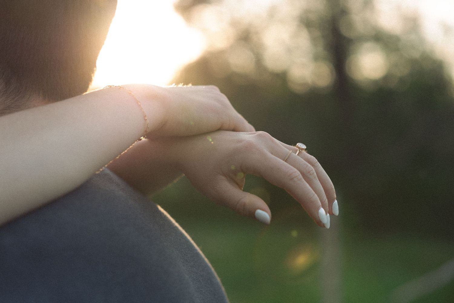 Close-up of a woman's hand with an engagement ring resting on a man's shoulder at sunset outdoors.