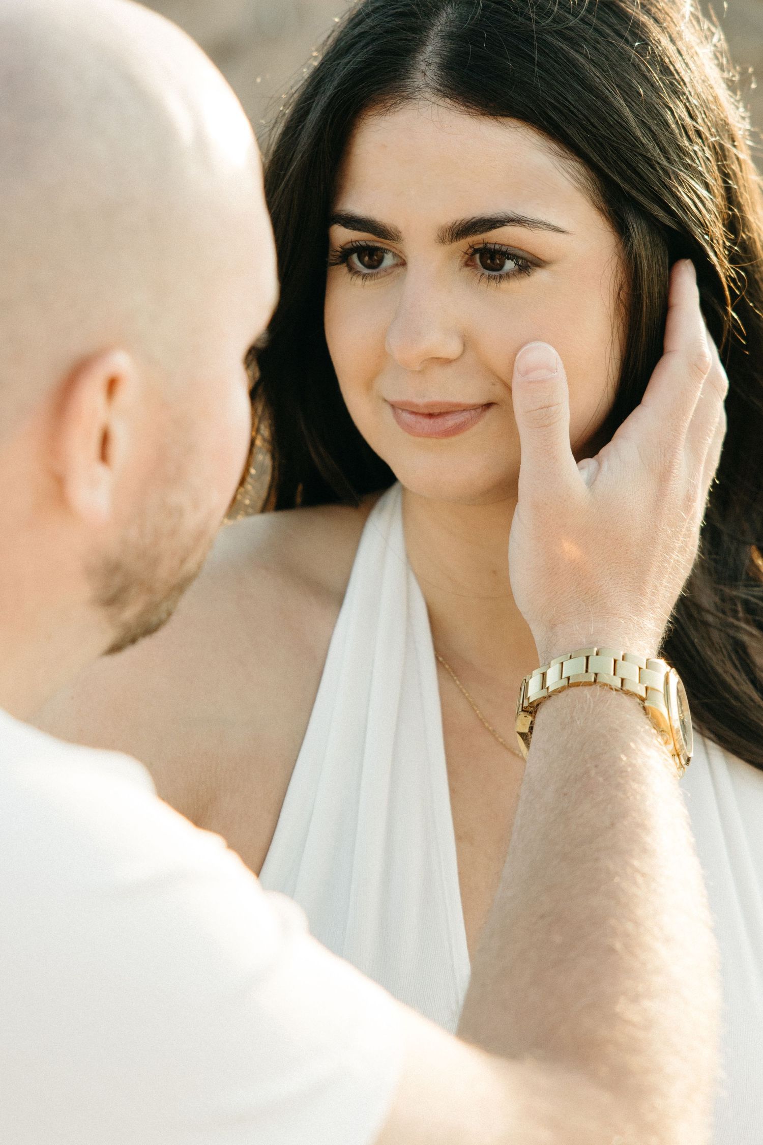 Man tenderly touching womans face as she gazes up at him during romantic beach engagement session.