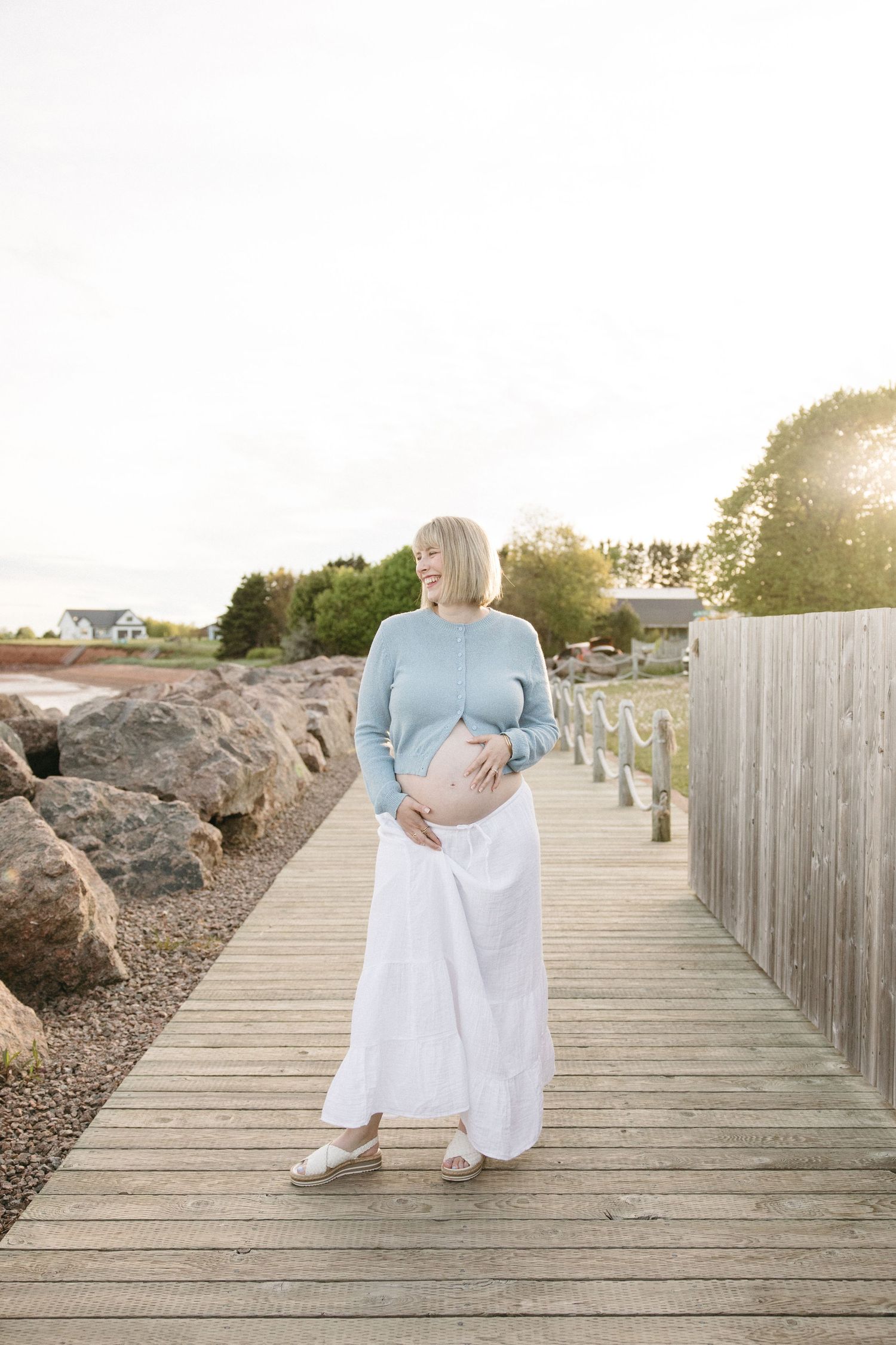 Pregnant woman in blue sweater and white skirt cradling her baby bump on a sunny wooden boardwalk by the ocean.