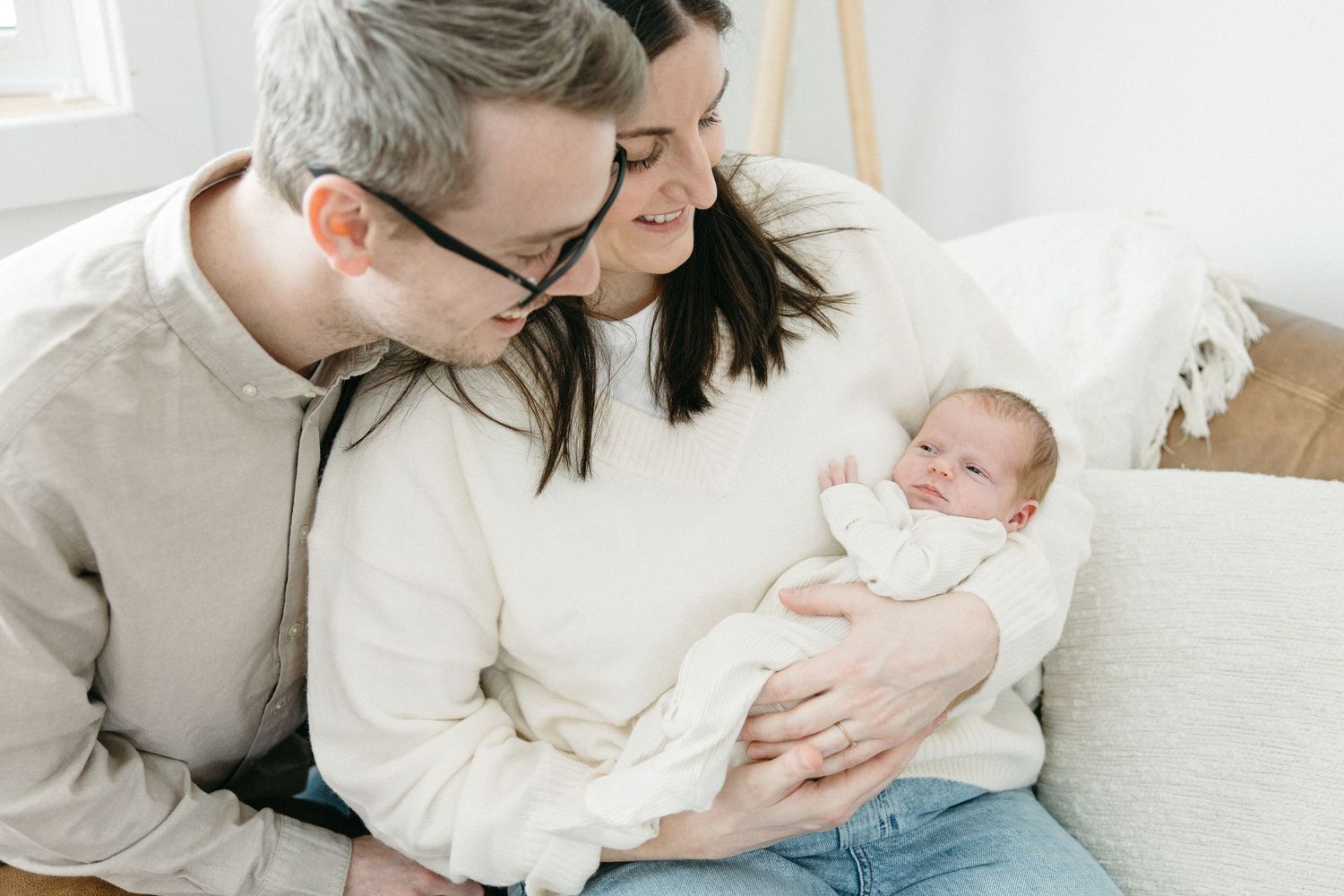 A parent in white clothing holds and lovingly gazes at their newborn baby while sitting on a light-colored couch.