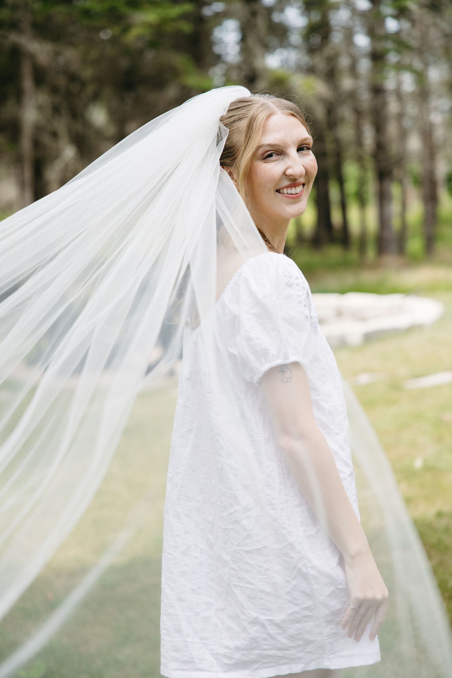 Smiling bride in white dress with flowing veil twirls outdoors in pine forest setting.