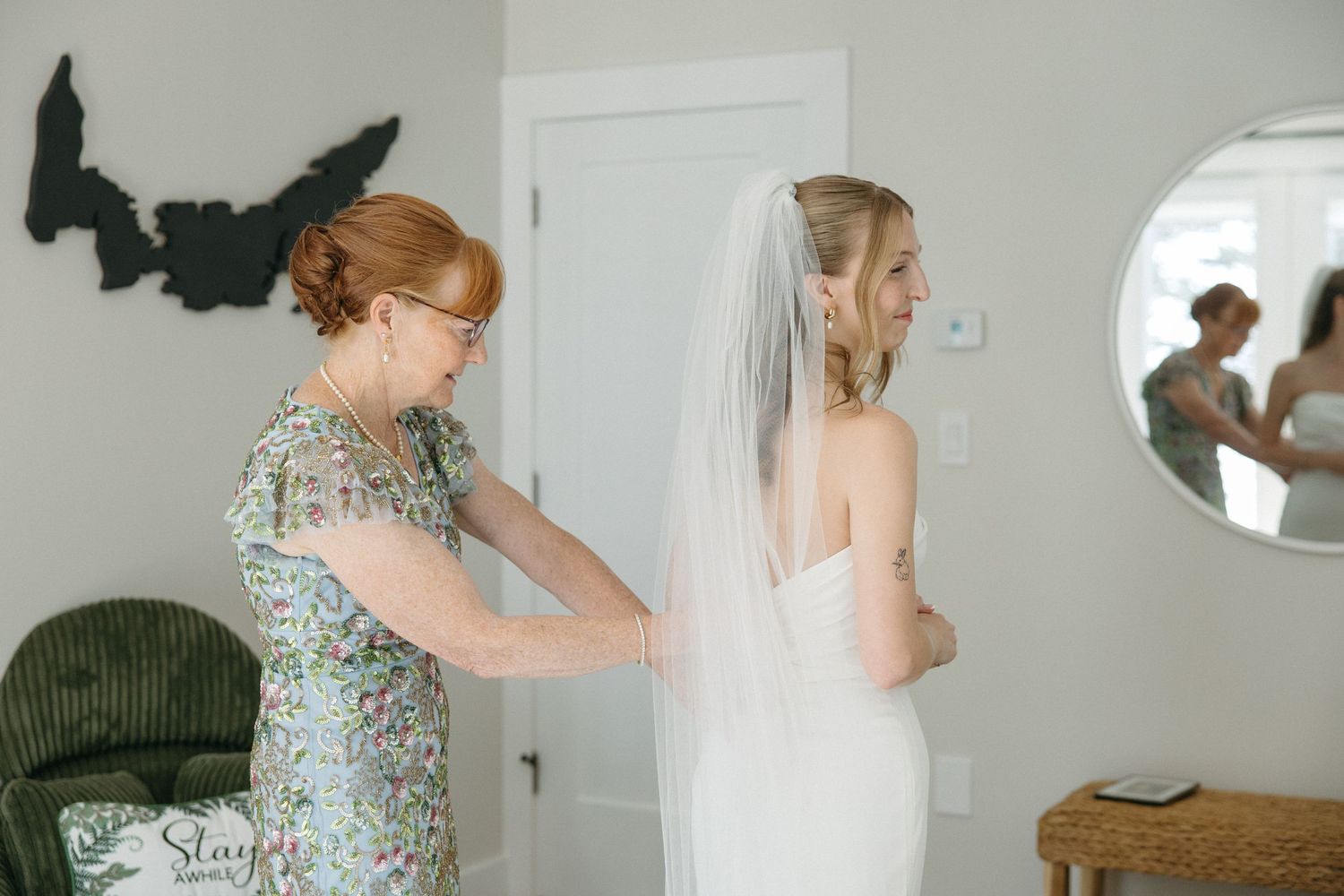 Mother helping bride with wedding dress zipper while bride wears veil, reflected in round mirror.