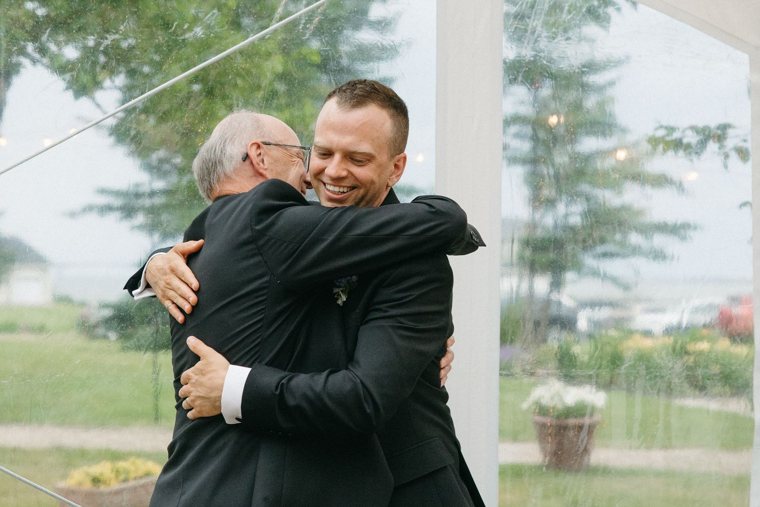 Two men share a warm embrace under a tent at a wedding celebration.