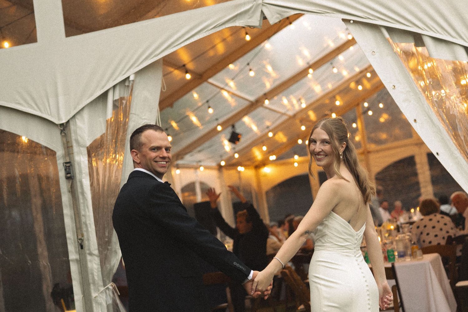 Smiling bride and groom hold hands as they enter their tented wedding reception with string lights overhead.