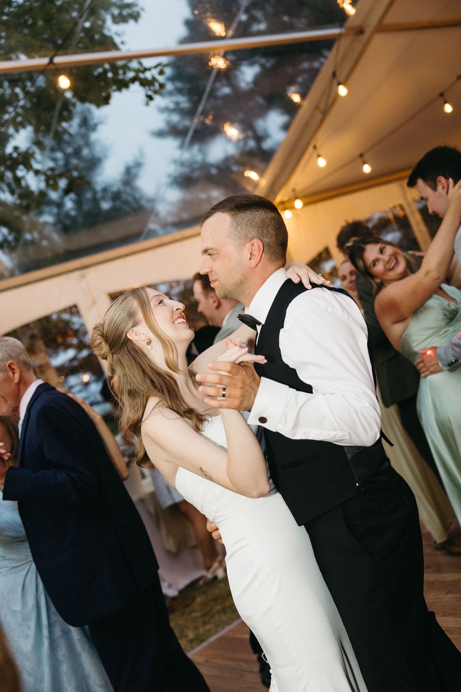 Bride and groom share a joyful first dance under string lights at their elegant tented wedding reception.