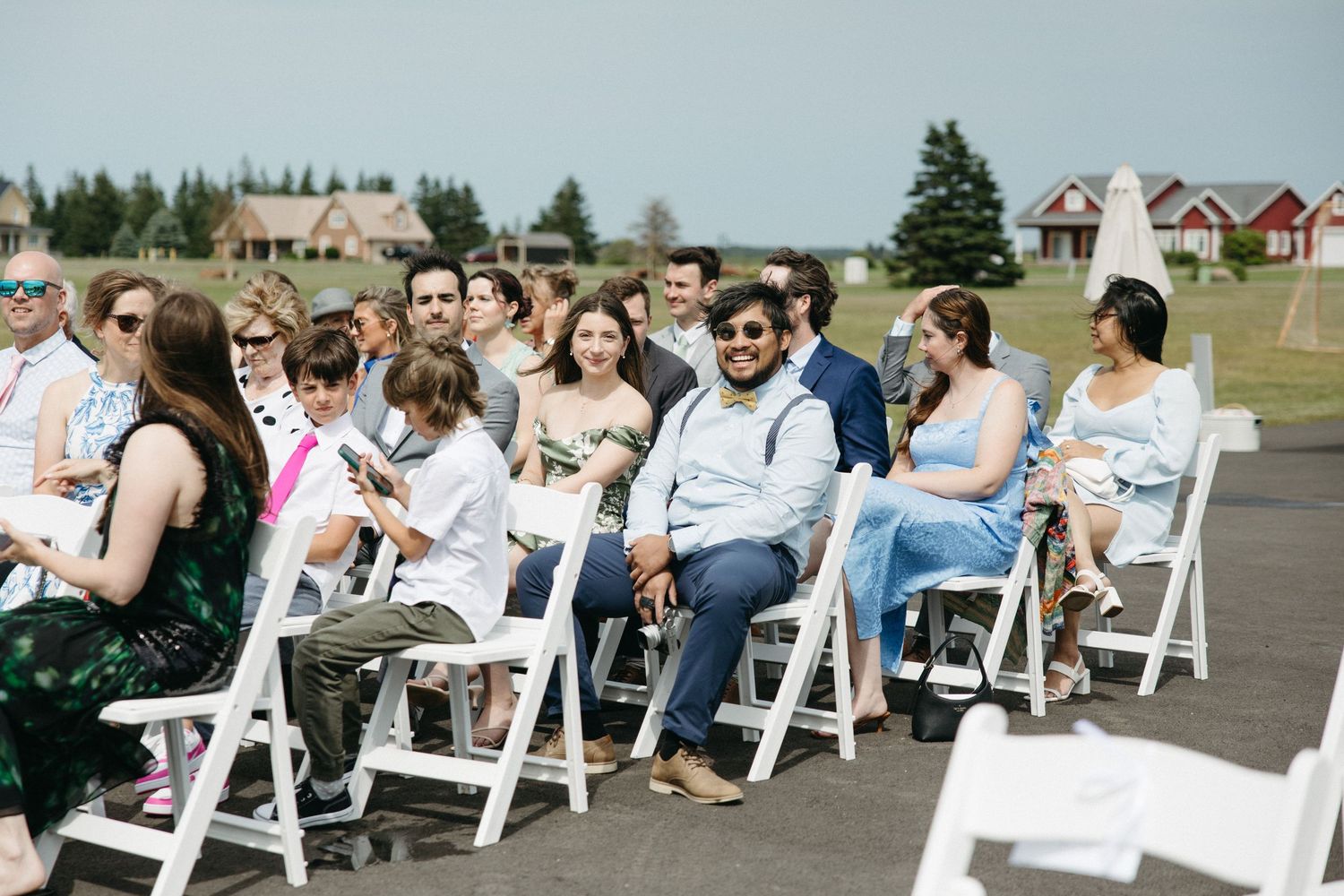 Wedding guests seated in white chairs outdoors during sunny ceremony.