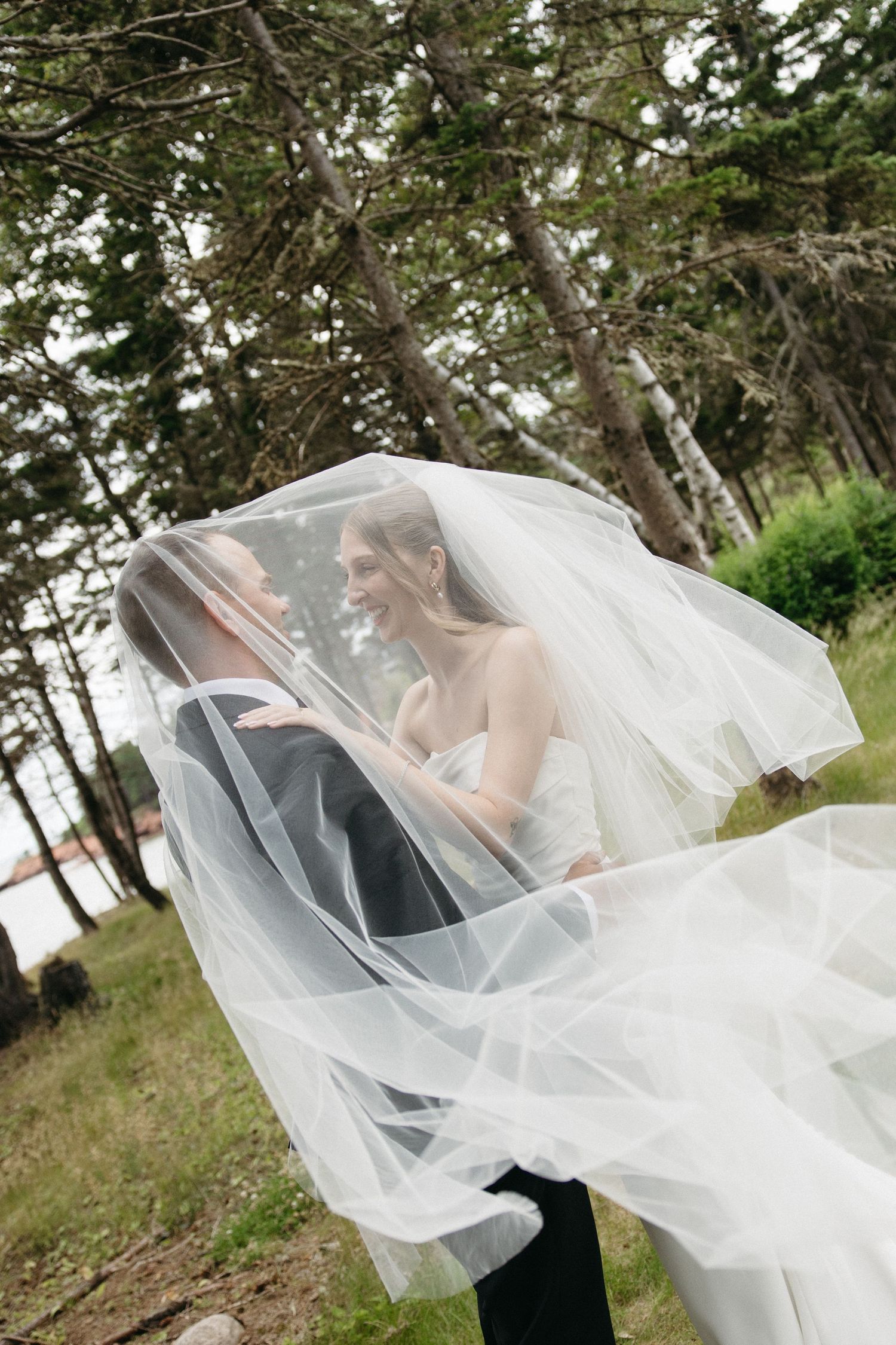 Bride and groom share a romantic moment hidden beneath a flowing white wedding veil in a forest setting.